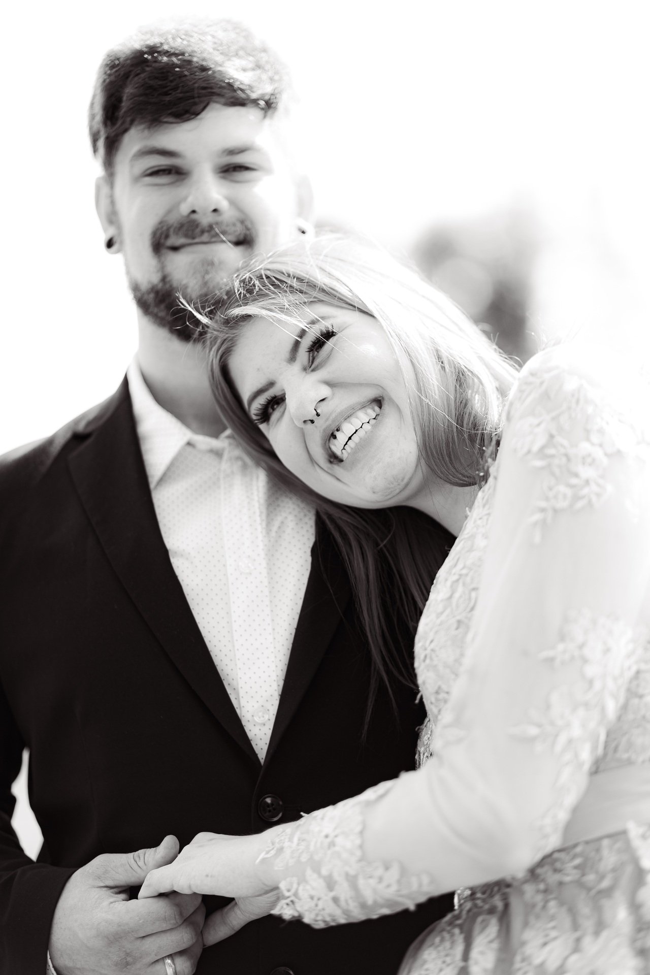 Schwarz-weiß-Foto eines jungen Paares, das glücklich lächelt. Die Frau lehnt ihren Kopf auf die Schulter des Mannes. Beide tragen elegante Kleidung, die Frau ein Spitzenkleid und der Mann einen Anzug.