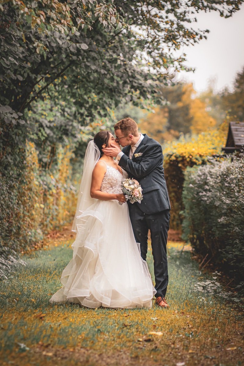 Hochzeitspaar küsst sich im Garten, Braut trägt ein weißes Kleid und Brautstrauß, Bräutigam im Anzug.
