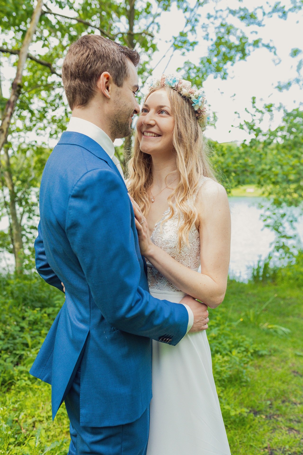 Ein Brautpaar steht im Freien in einem grünen, bewaldeten Gebiet. Die Braut trägt ein weißes Kleid und einen Blumenkranz im Haar. Der Bräutigam trägt einen blauen Anzug. Sie schauen sich lächelnd in die Augen.