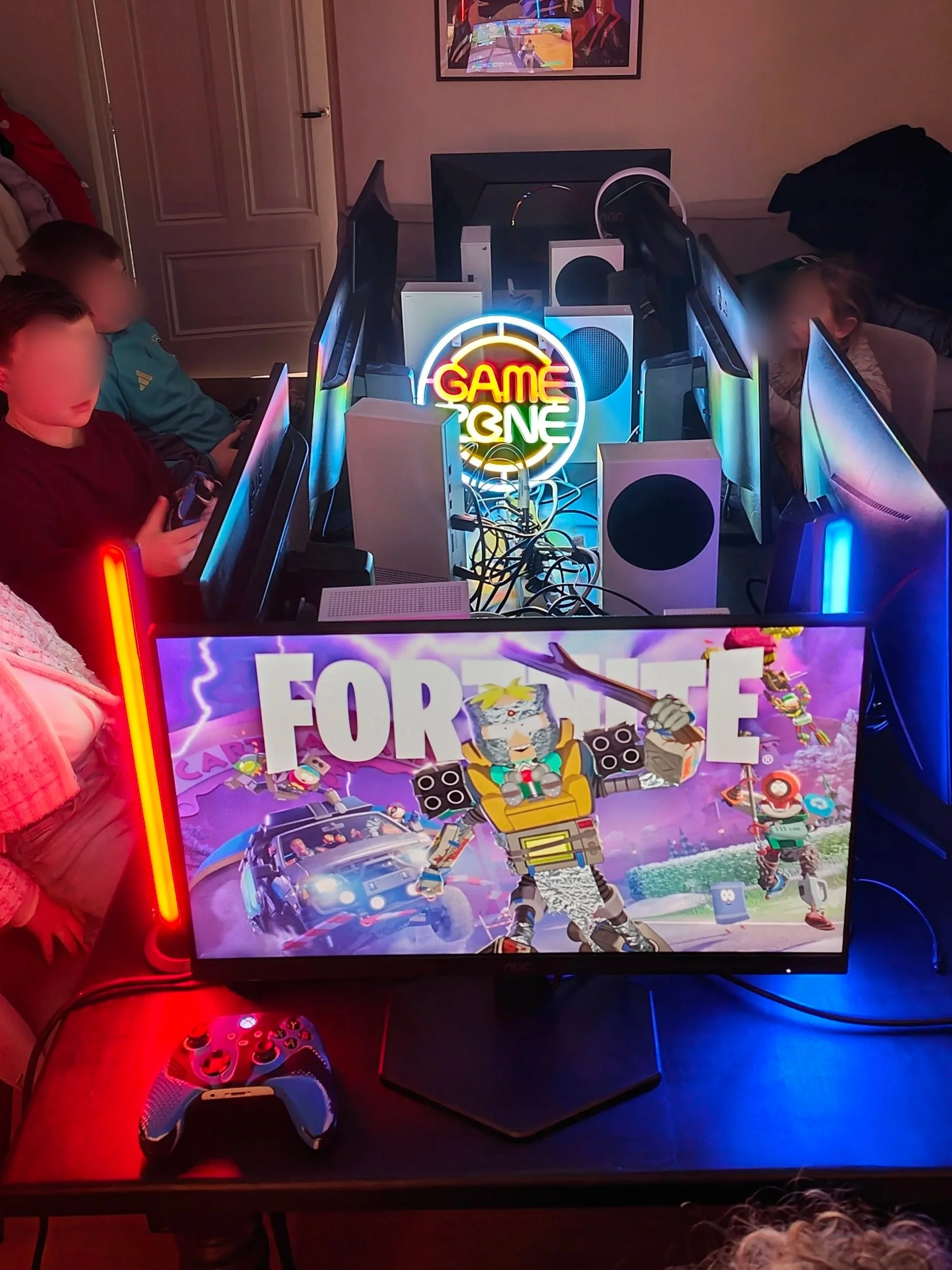 Gezeten kinderen die gamen op meerdere computers met een neonlicht dat zegt 'GAME ON'. In het midden op het scherm, het spel 'Fortnite' met een kleurrijk achtergrond.
