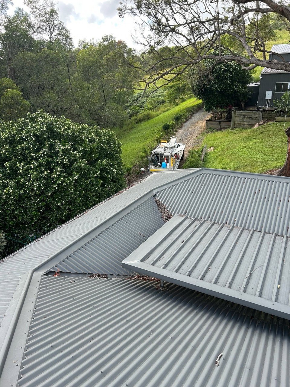 View of a corrugated metal roof with a section lifted to reveal debris underneath, with a landscape of trees and a trailer parked on a dirt path in the background.