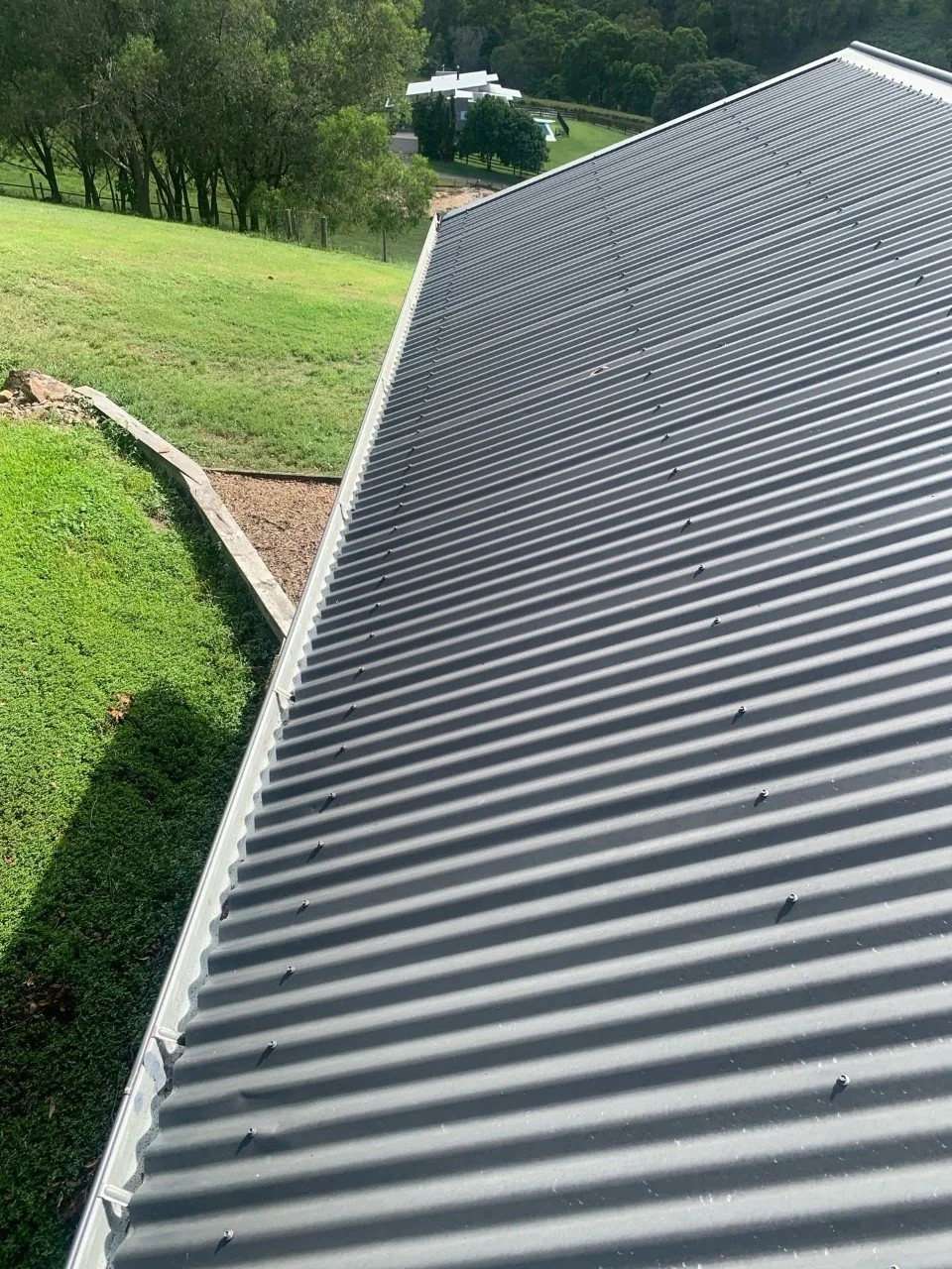 Close-up of a corrugated metal roof with shadows cast from the sun, with green grassy slope and trees in the background.