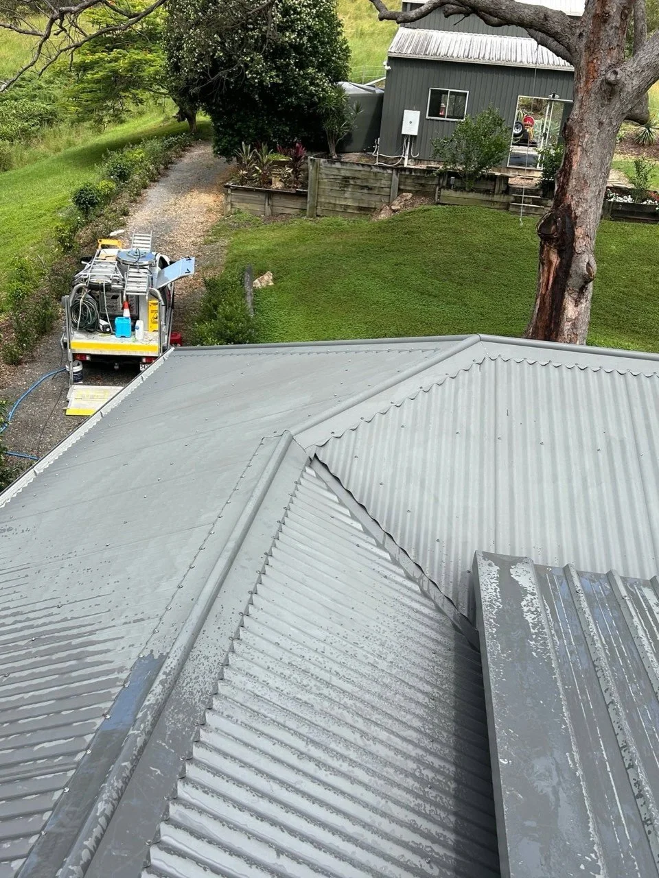 View from a roof showing a metal roof with gutters, a large tree, a grassy yard, a gravel pathway, and a gray house in the background with some outdoor furniture and plants.