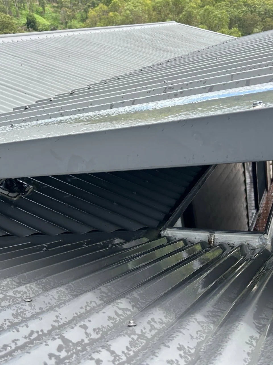 Close-up view of metal roofing panels with rainwater on the surface and a window opening in the building.