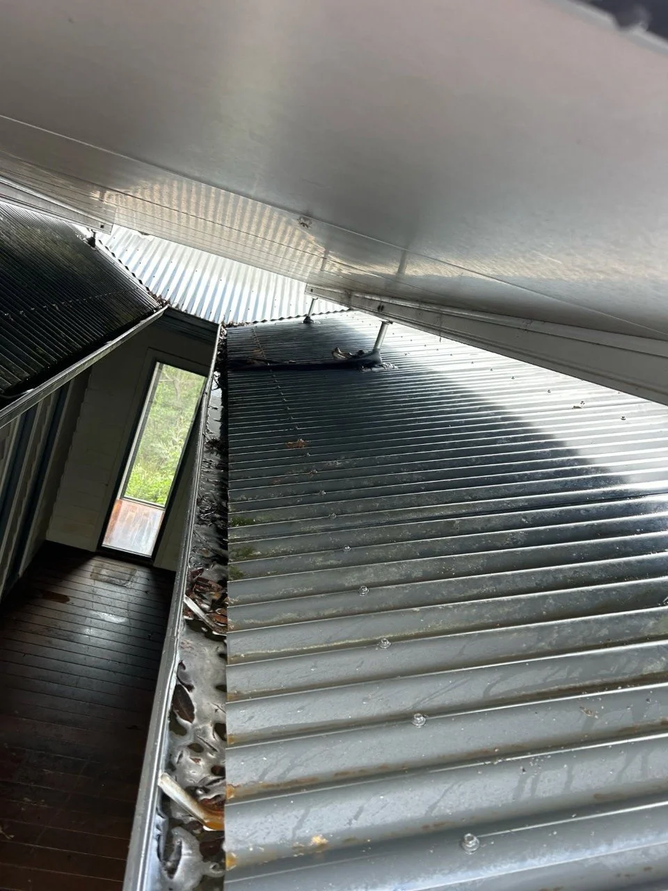 View of a rooftop with metal sheets, showing signs of damage along the edges and around fasteners, next to an interior room with hardwood floor and a glass door leading outside.