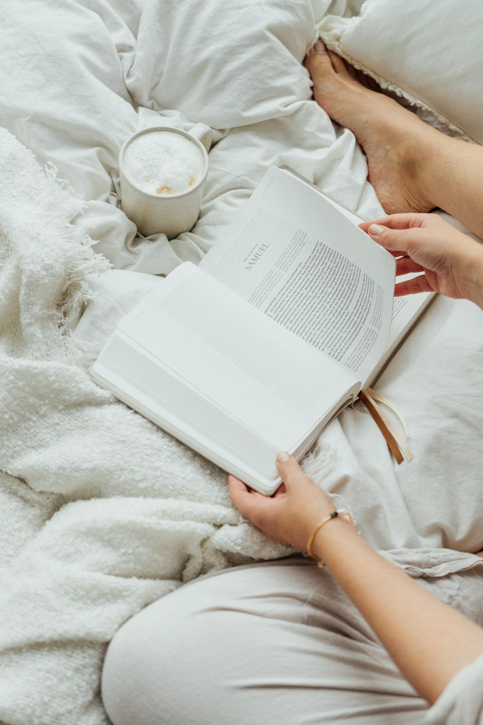 A person lying in bed, wearing light gray pajama pants, holding an open book, with a white mug of foamy coffee or tea, a white mattress, and a white blanket.