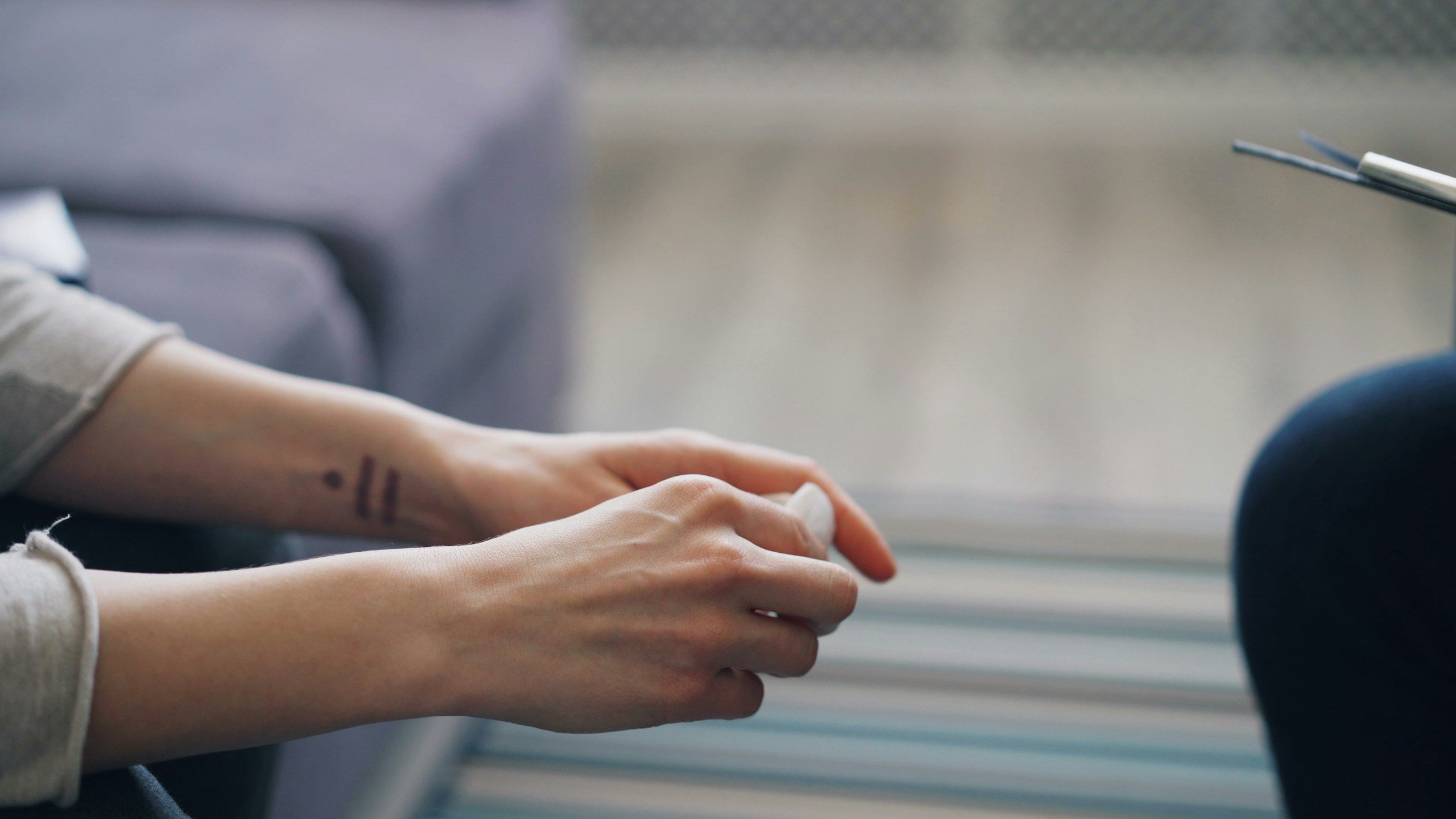 Close-up of a person's hands, with a tattoo on the wrist, resting on their lap, in a setting with a blurred background and a pair of chopsticks visible to the right.