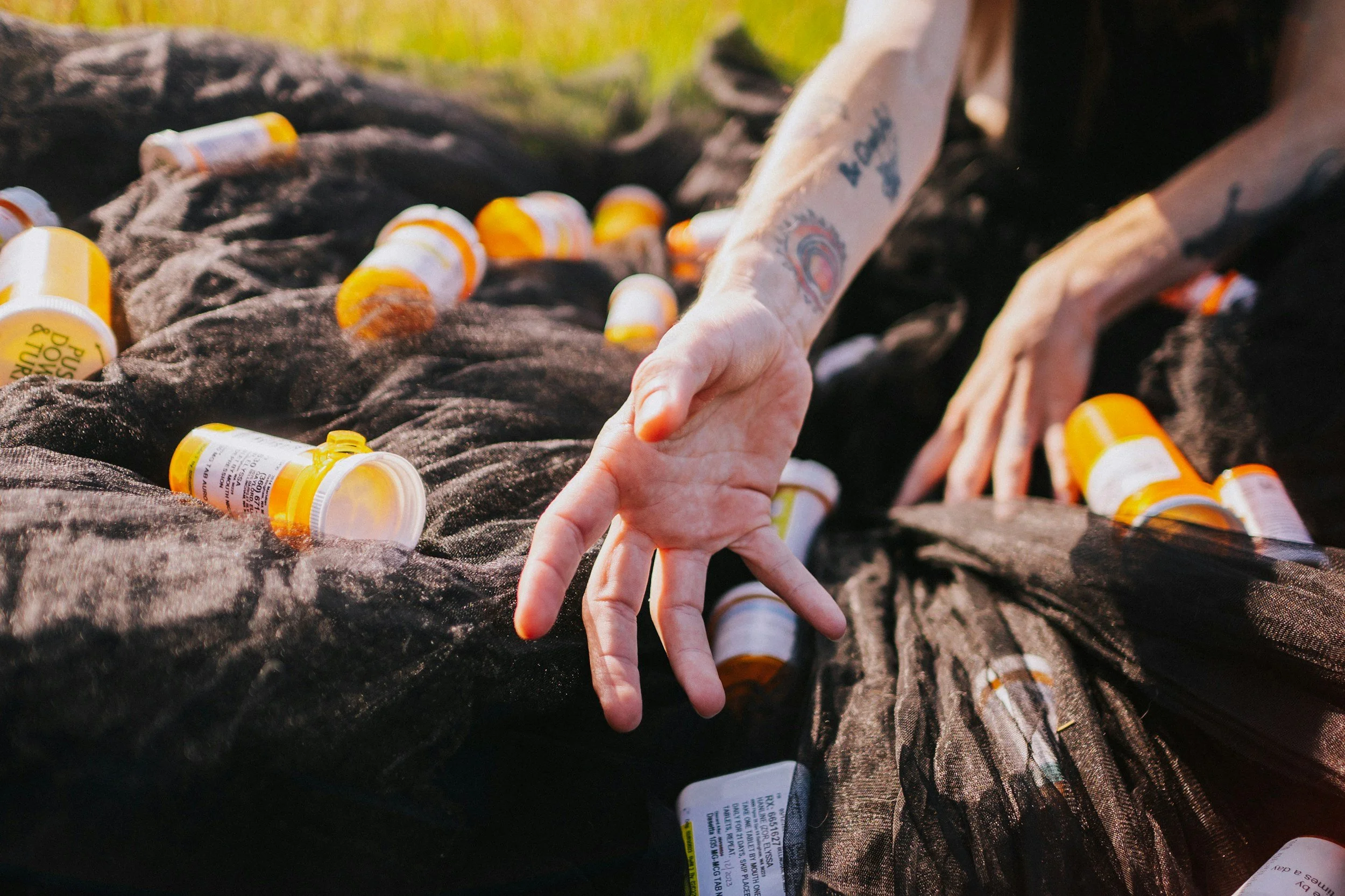 Close-up of hands with tattoos reaching over black fabric with numerous prescription pill bottles scattered on it outdoors.