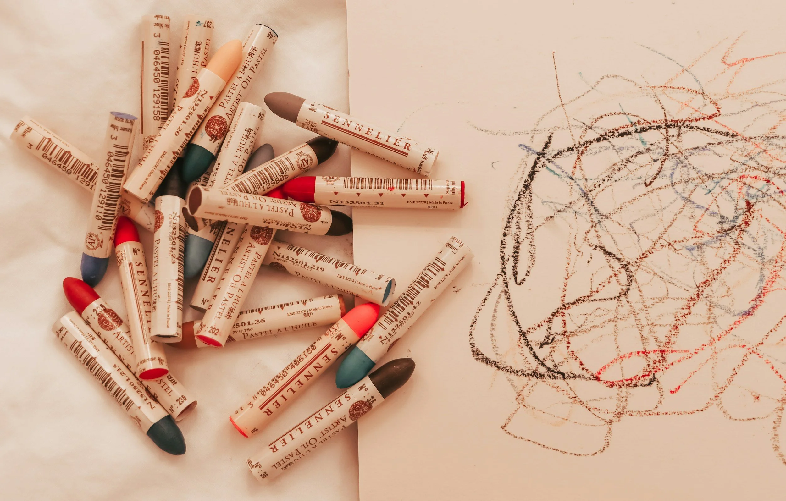 A pile of Crayola model Oil Pastels with various colors on a white surface, alongside a child's drawing of a star and some scribbles made with the pastels.