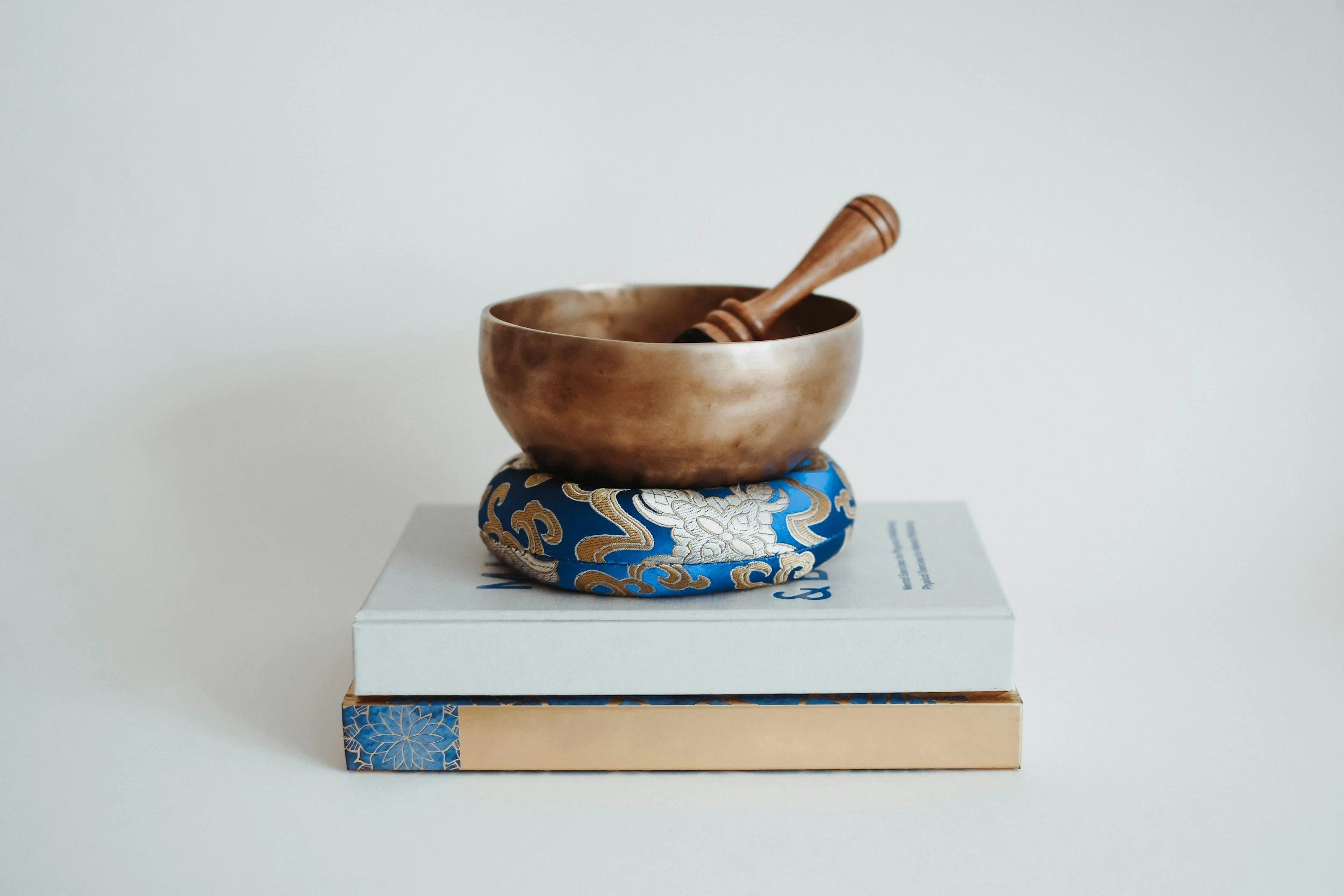 A brass singing bowl with a wooden mallet resting inside, placed on a decorative base with blue and gold patterns, on top of a white box.