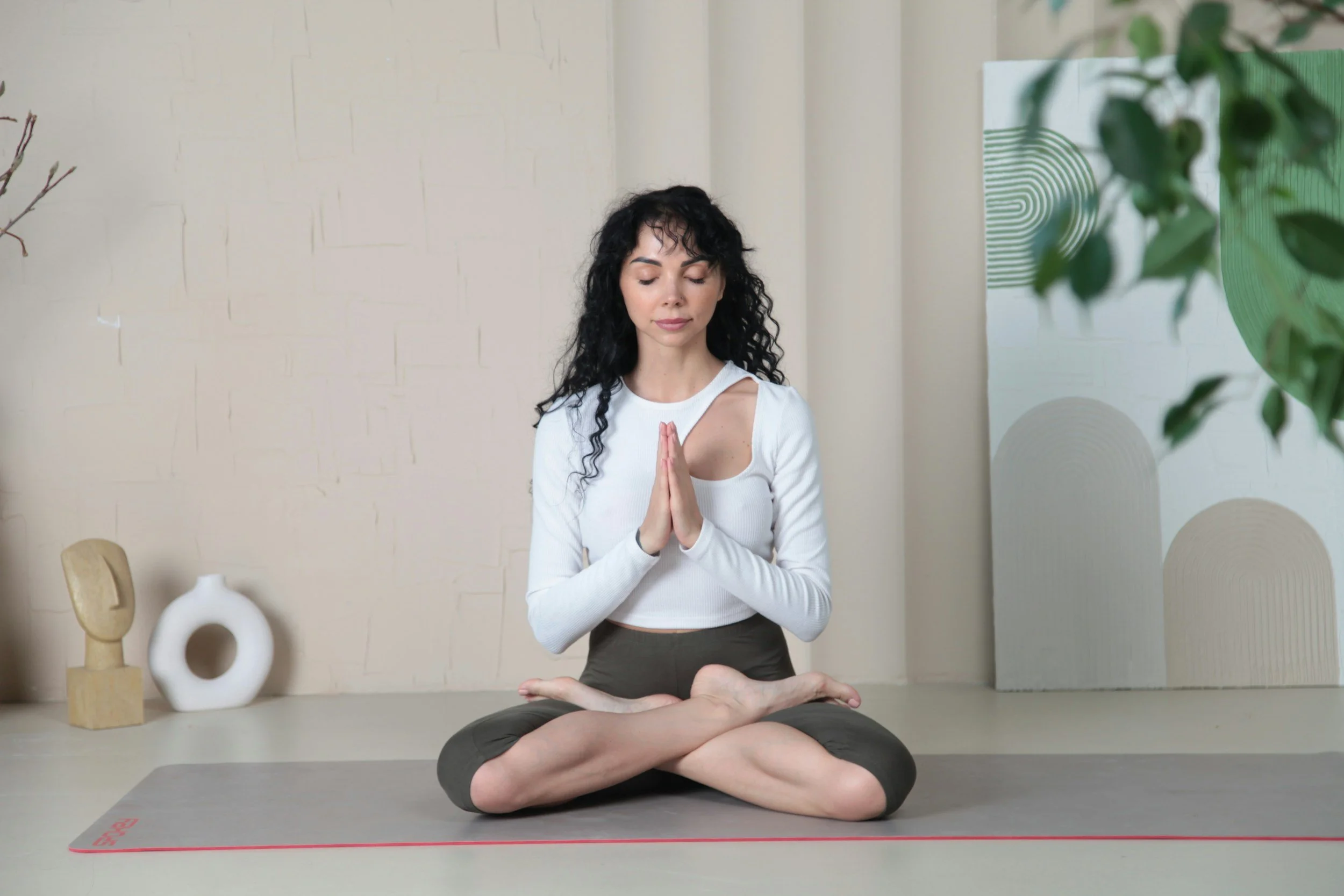 A woman practicing yoga indoors, seated on a yoga mat with her legs crossed and hands pressed together in prayer position, with a person lying under her in a supported pose.