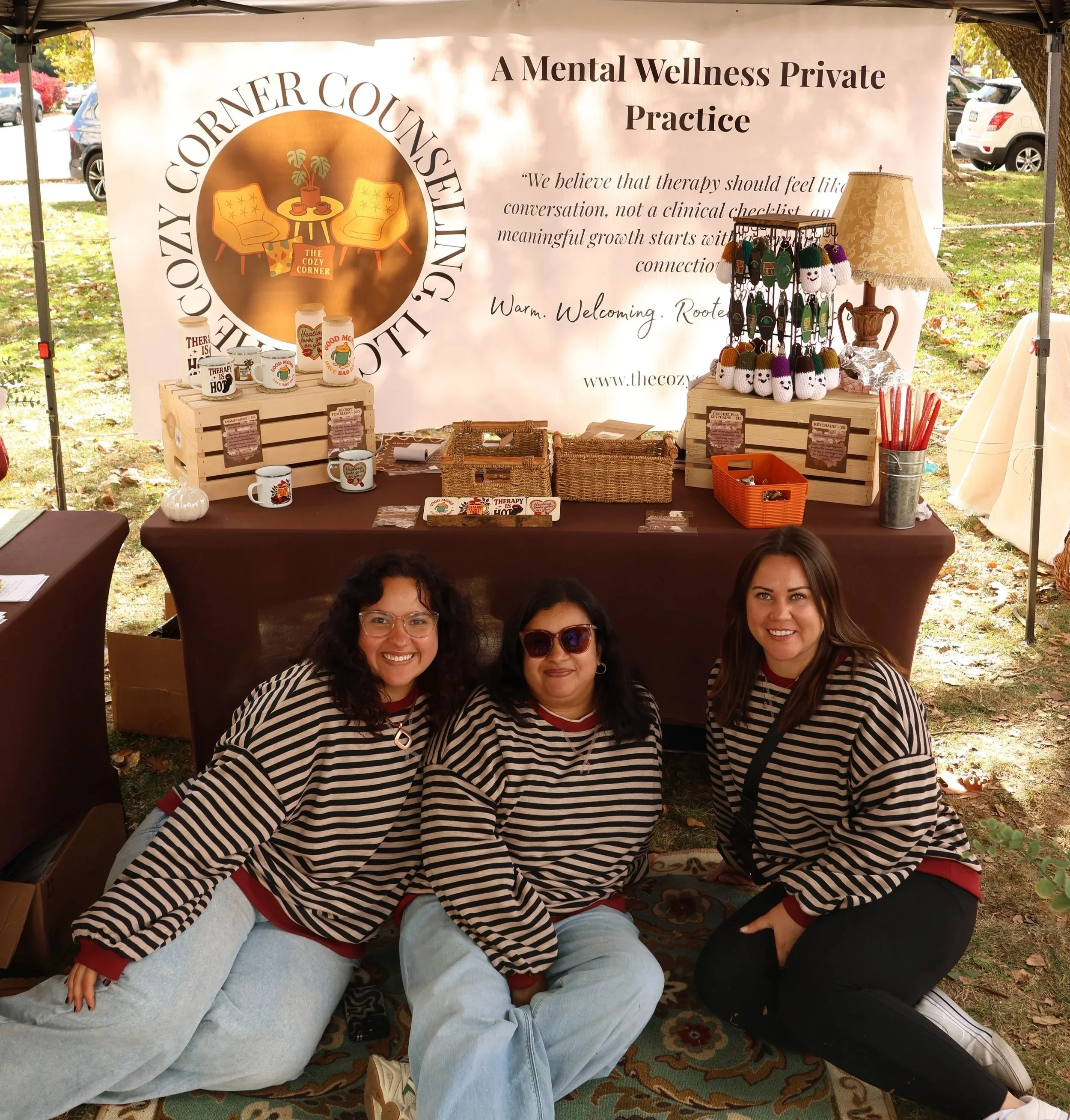 Three women in striped shirts posing in front of a booth at an outdoor event. The booth has a large banner reading 'A Mental Wellness Private Practice' and 'The Cozy Corner Counseling LLC,' with various merchandise on display, including mugs and handmade crochet items.