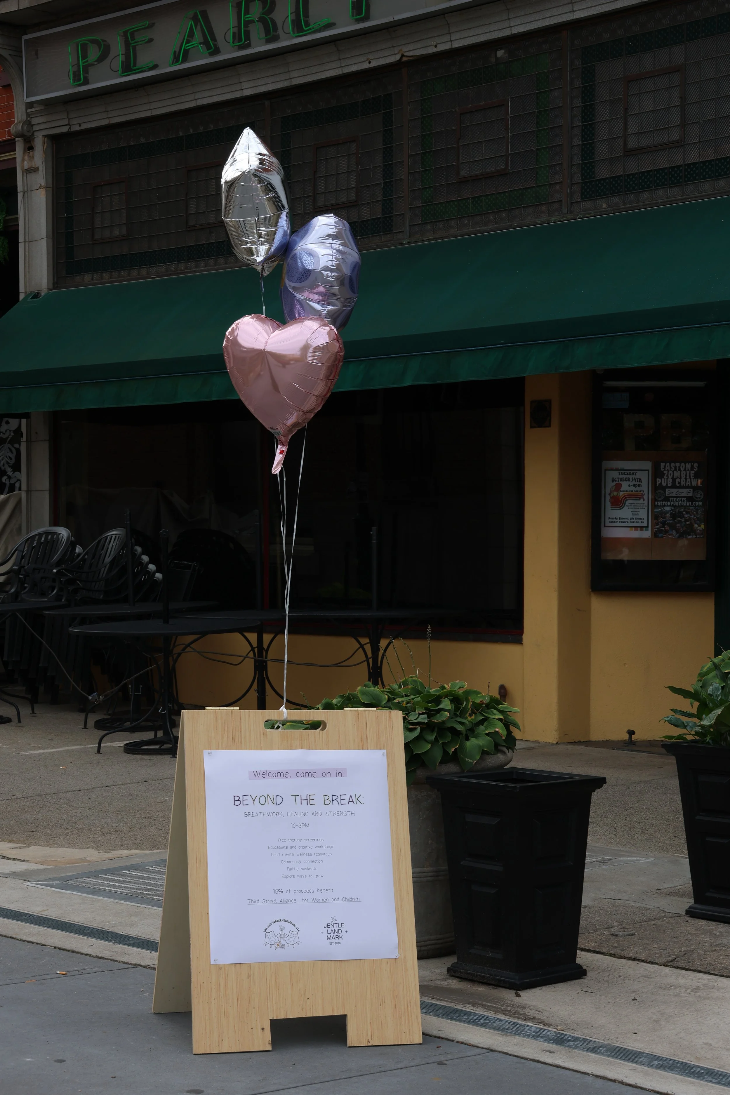 Three metallic balloons in silver, purple, and pink attached to a wooden sidewalk sign outside a cafe or restaurant.