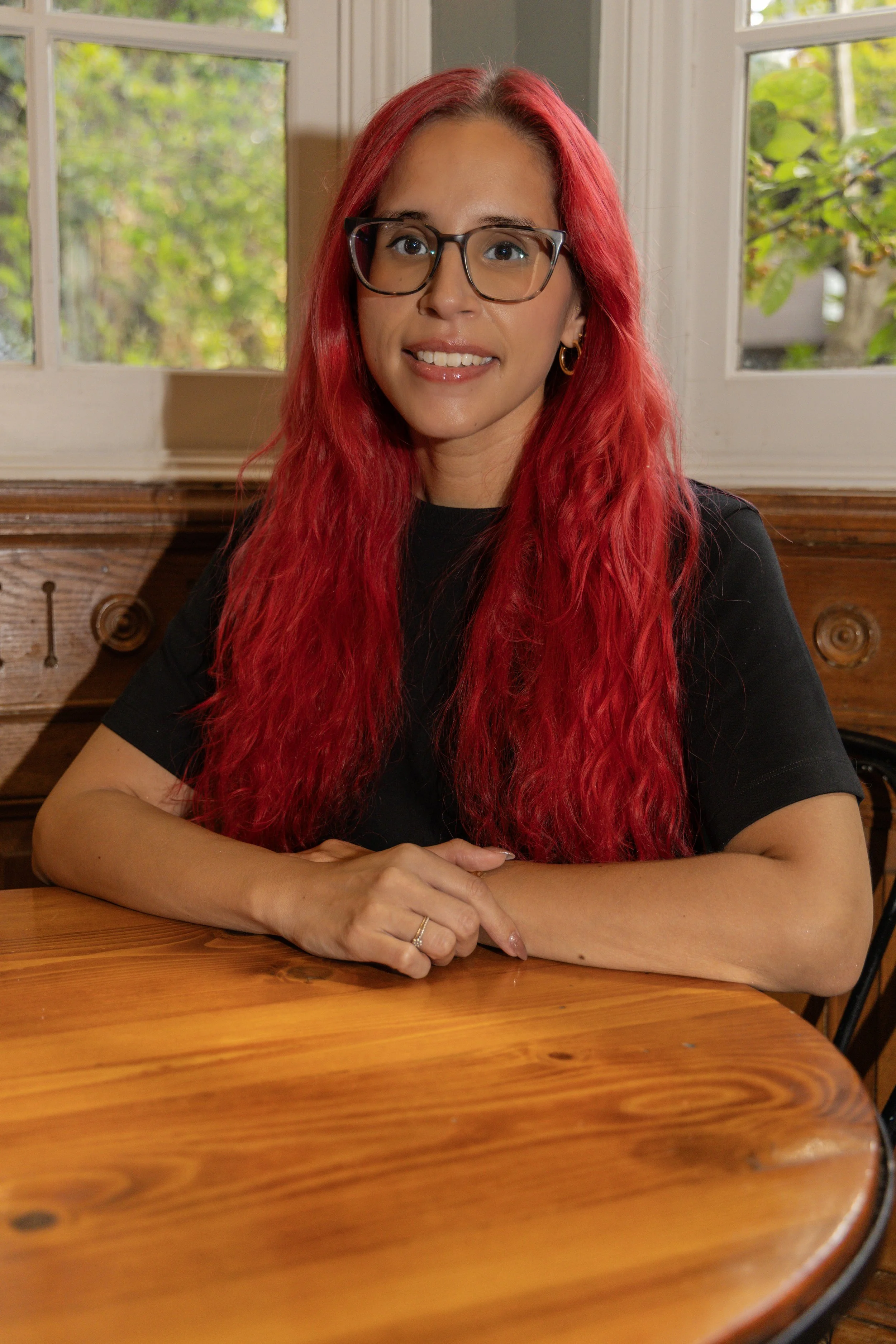 A woman with bright pink hair and glasses sitting outdoors on a concrete surface, wearing a black t-shirt and blue jeans, smiling at the camera.