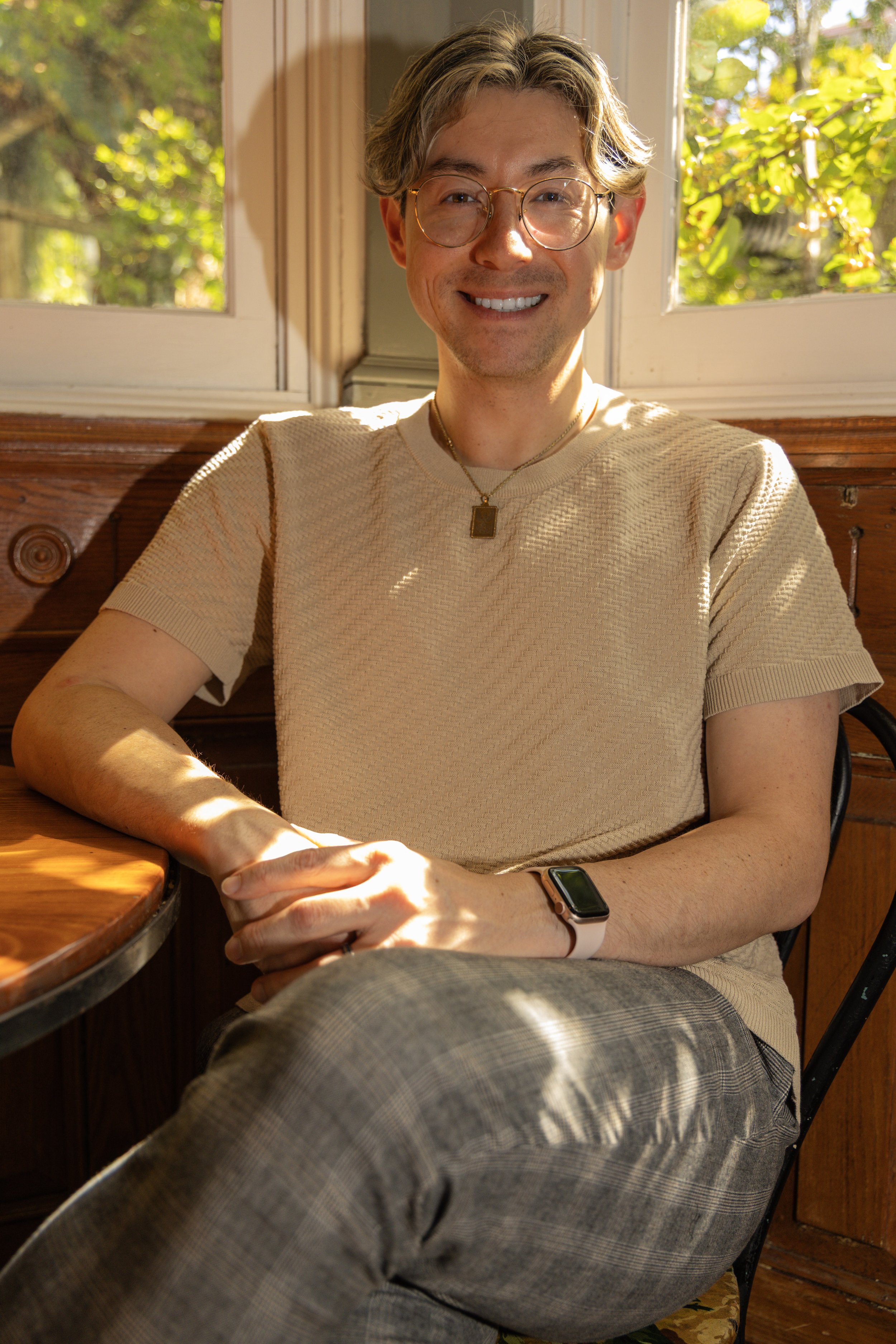 A smiling young man with glasses, short wavy hair, wearing a black shirt, sitting on a gray sofa with his hands clasped.