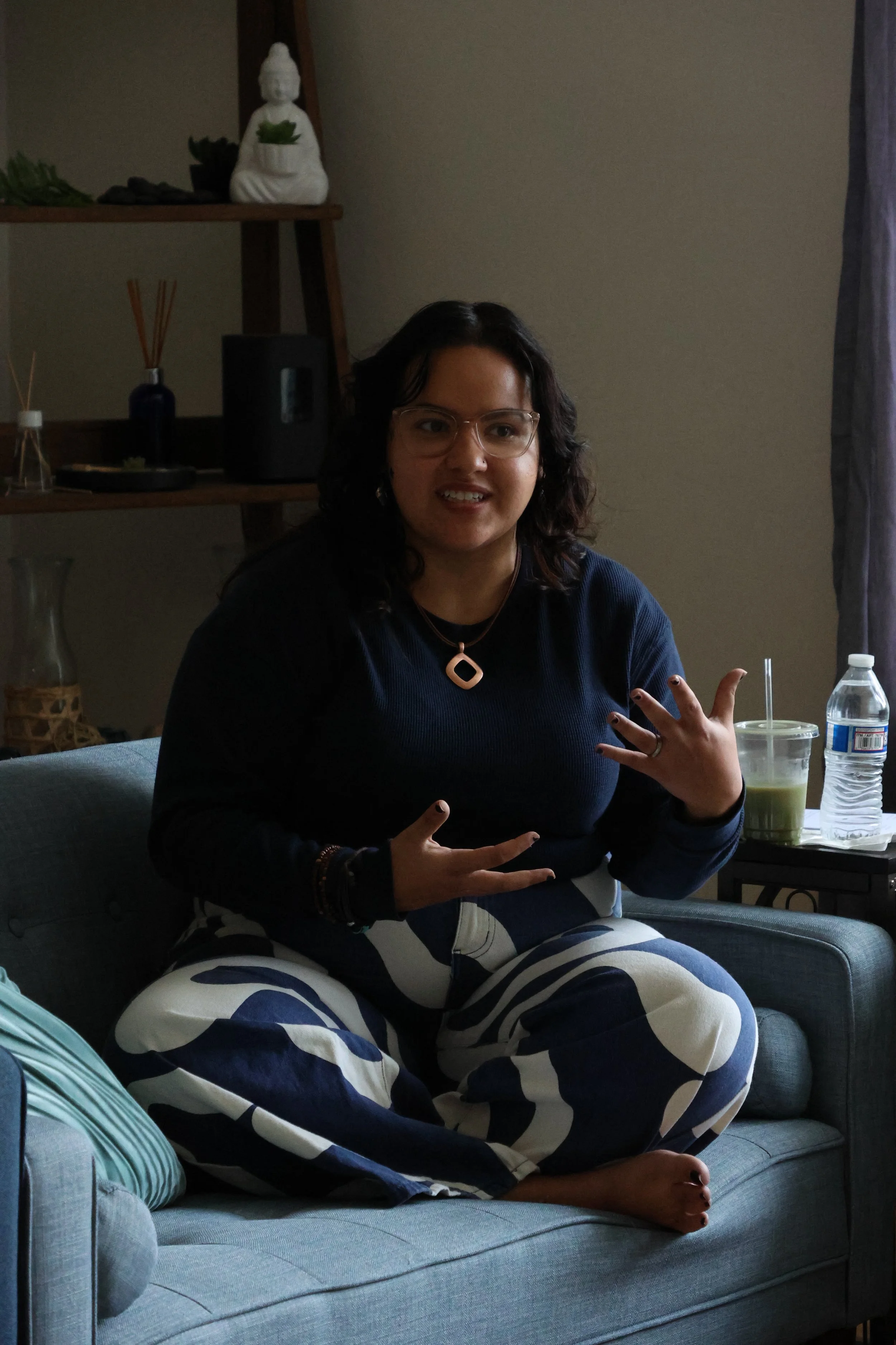 A woman with dark, curly hair, glasses, and jewelry, sitting on a blue sofa, cross-legged, making a hand sign, with glasses, a black shirt, and patterned pants, in a cozy room with a shelf and a window in the background.