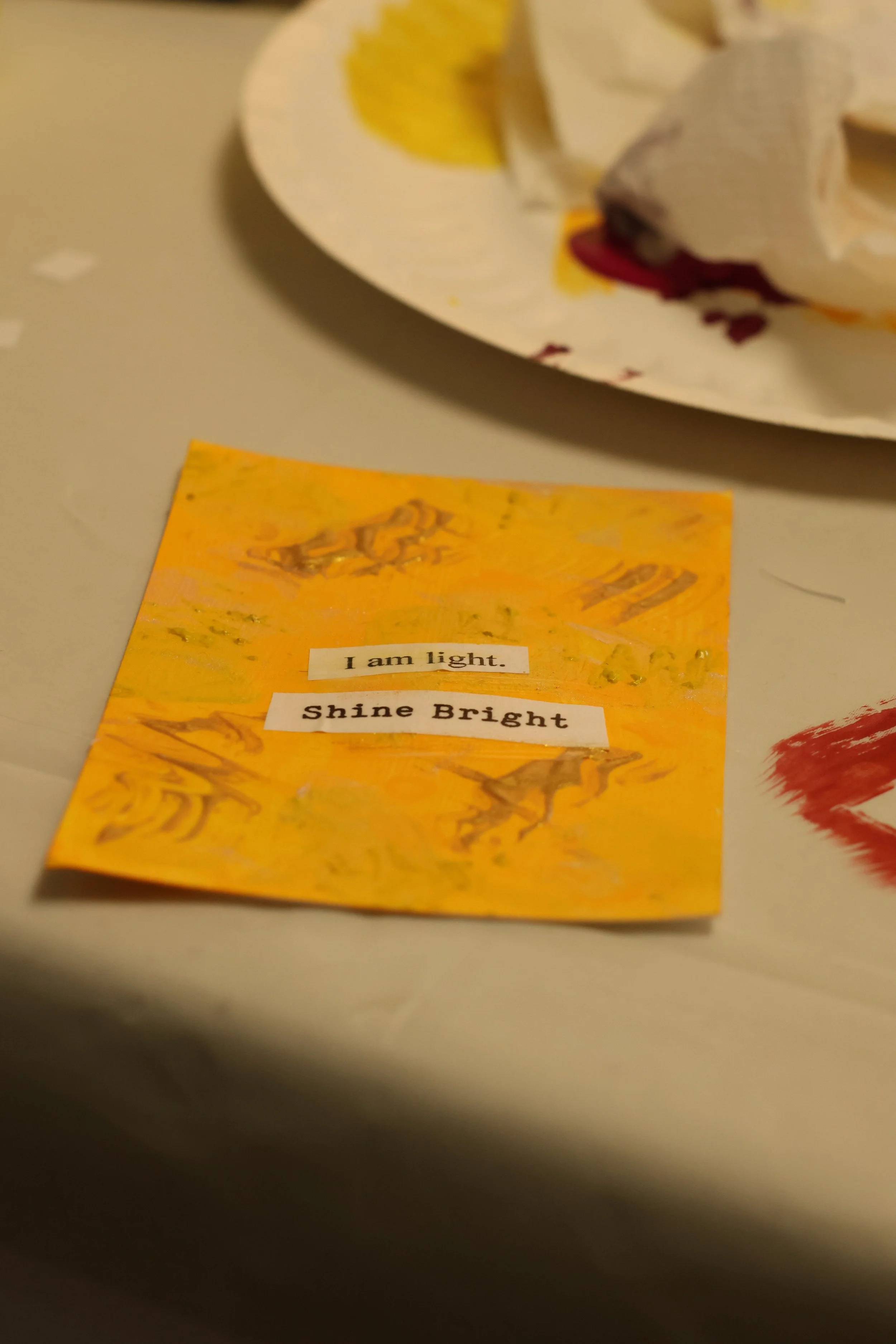 A small, orange, tie-dye piece of paper with the words 'I am light. Shine Bright' cut out and glued on. In the background, part of a paper plate with a crumpled paper napkin on it is visible.