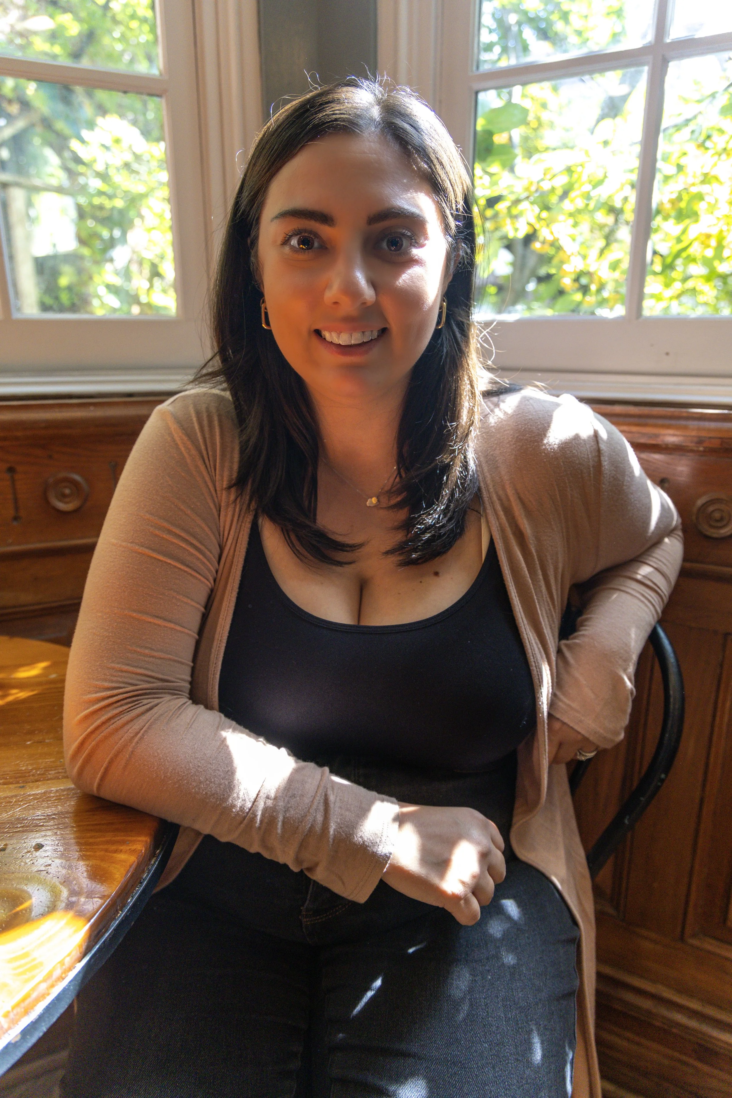 A woman with dark hair and brown eyes smiling, wearing gold hoop earrings and a black top, sitting in front of a large green leafy plant.