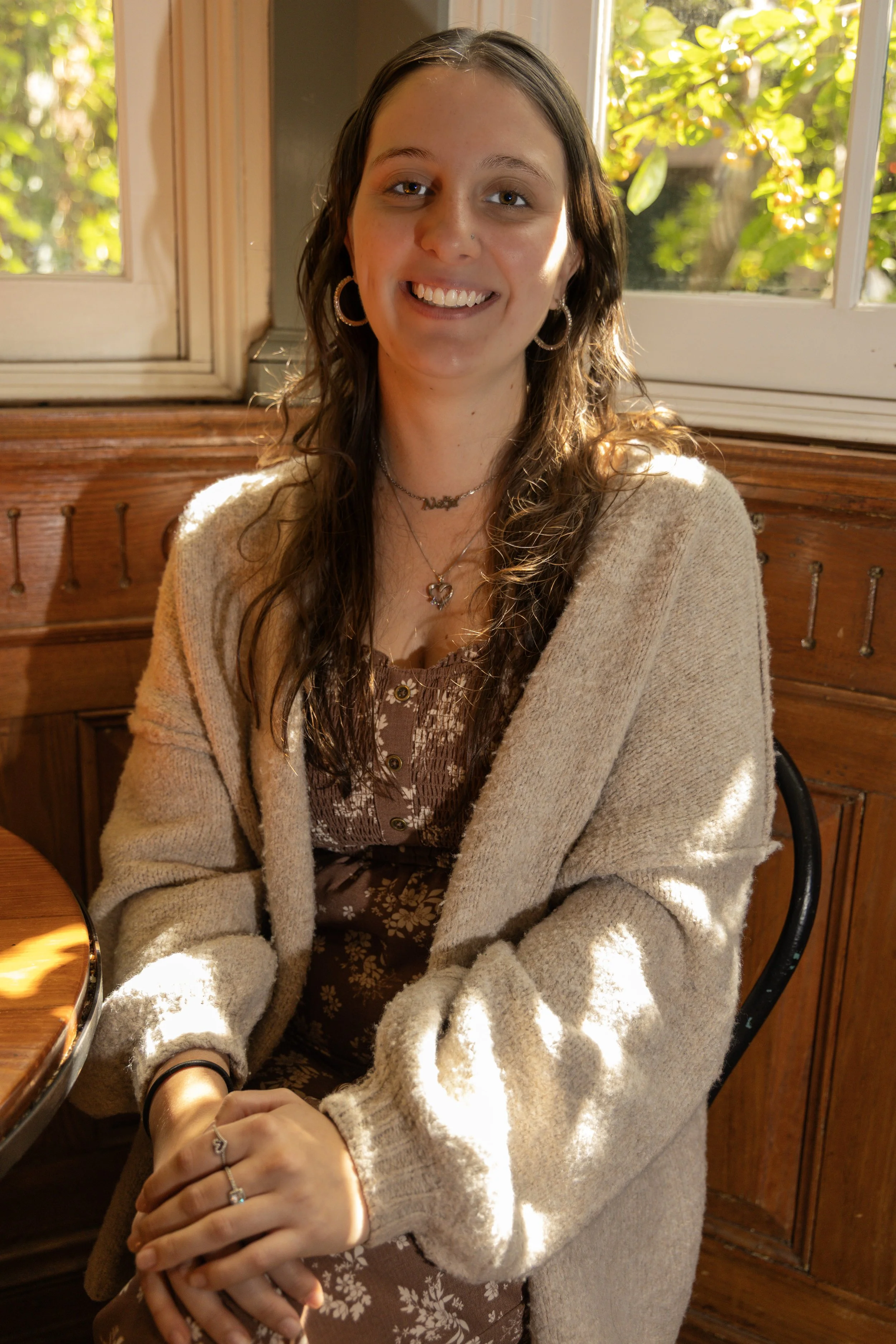 A young woman with long dark hair, wearing hoop earrings and a heart-shaped necklace, smiling outdoors in front of a flowering tree.