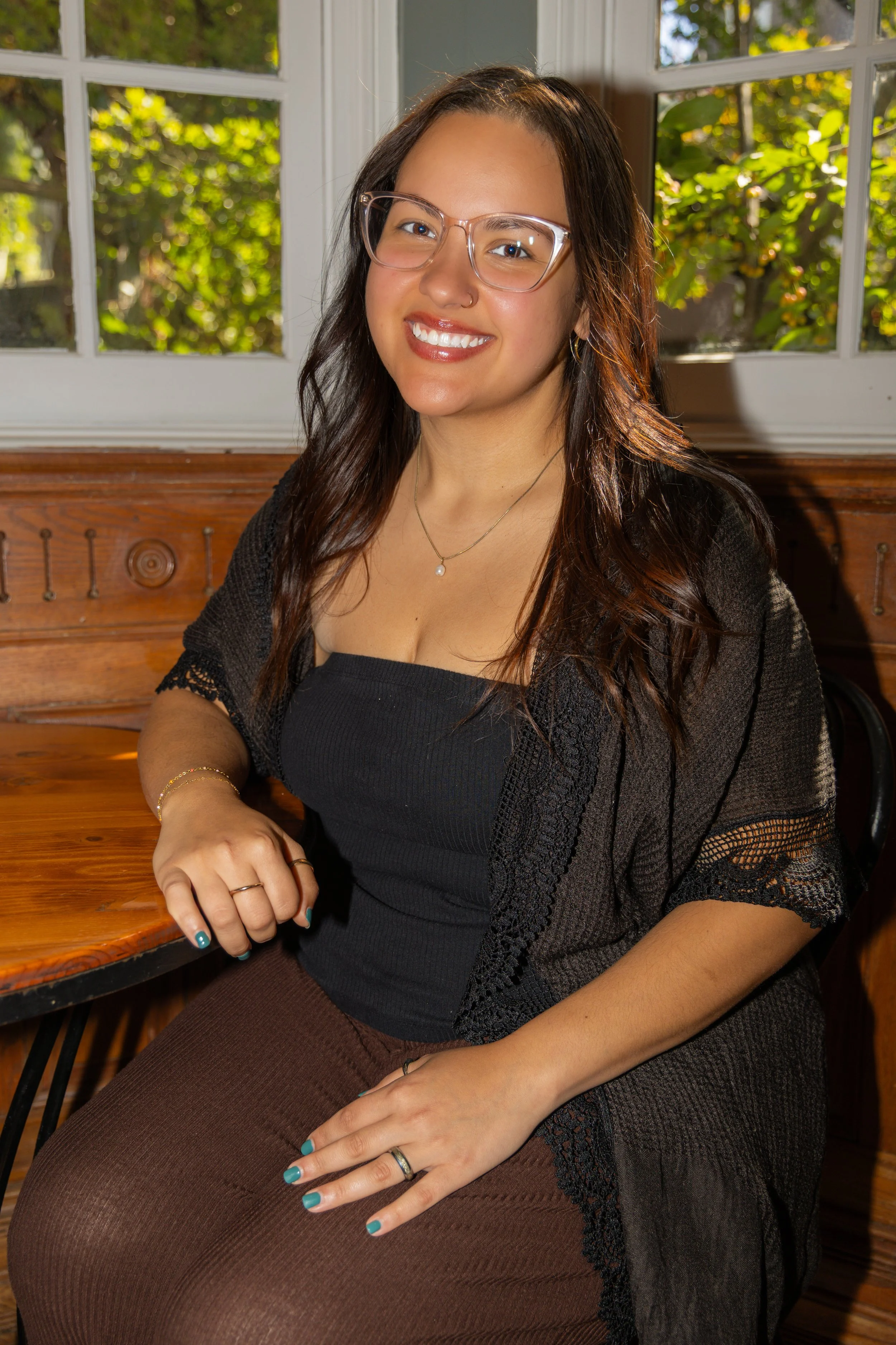 A woman with dark, wavy hair wearing a black top and a black shawl, smiling in front of a white brick wall.