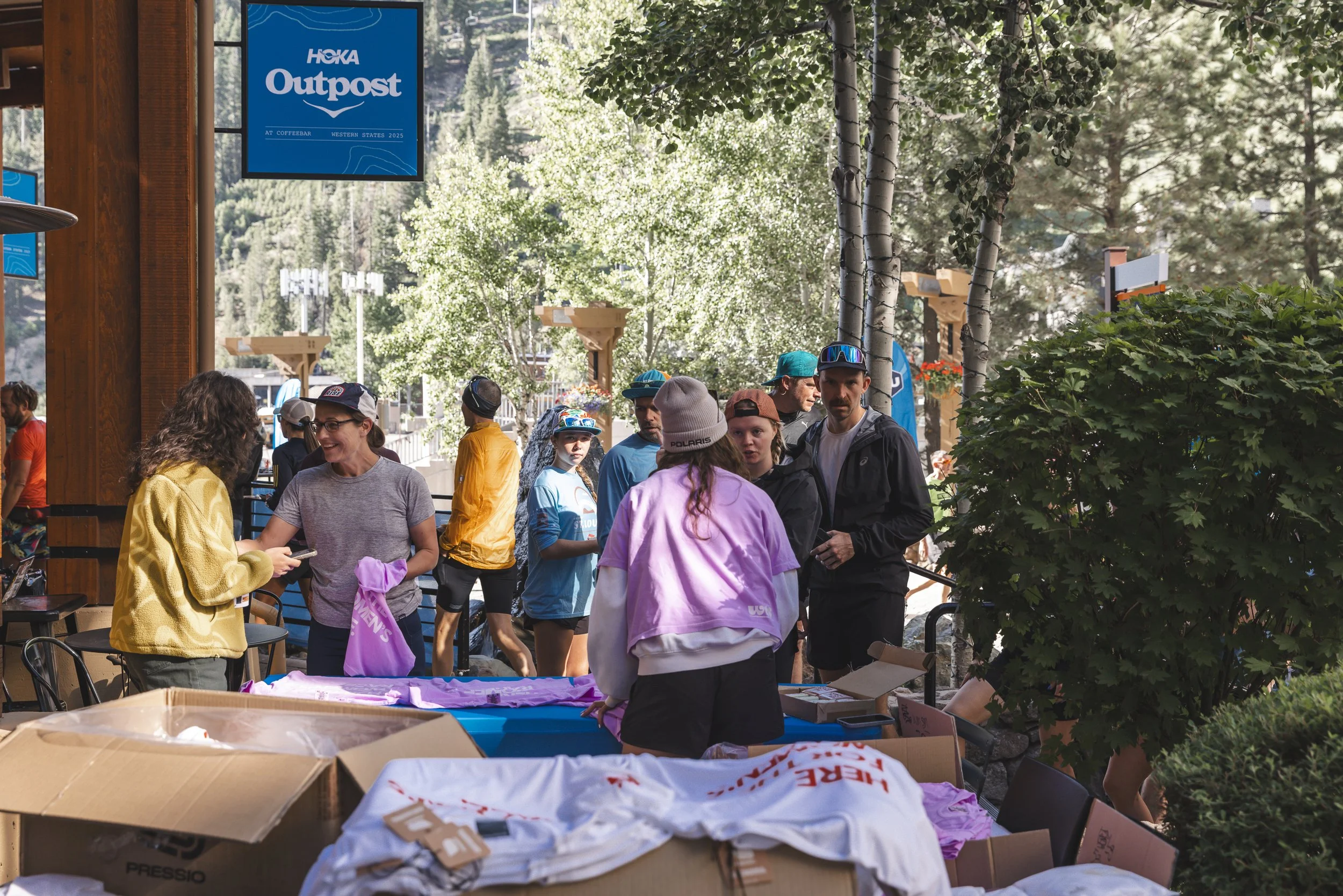 People gathered outside at the HOKA Outpost, with some browsing merchandise on a table and others in line, surrounded by trees and a mountain backdrop.