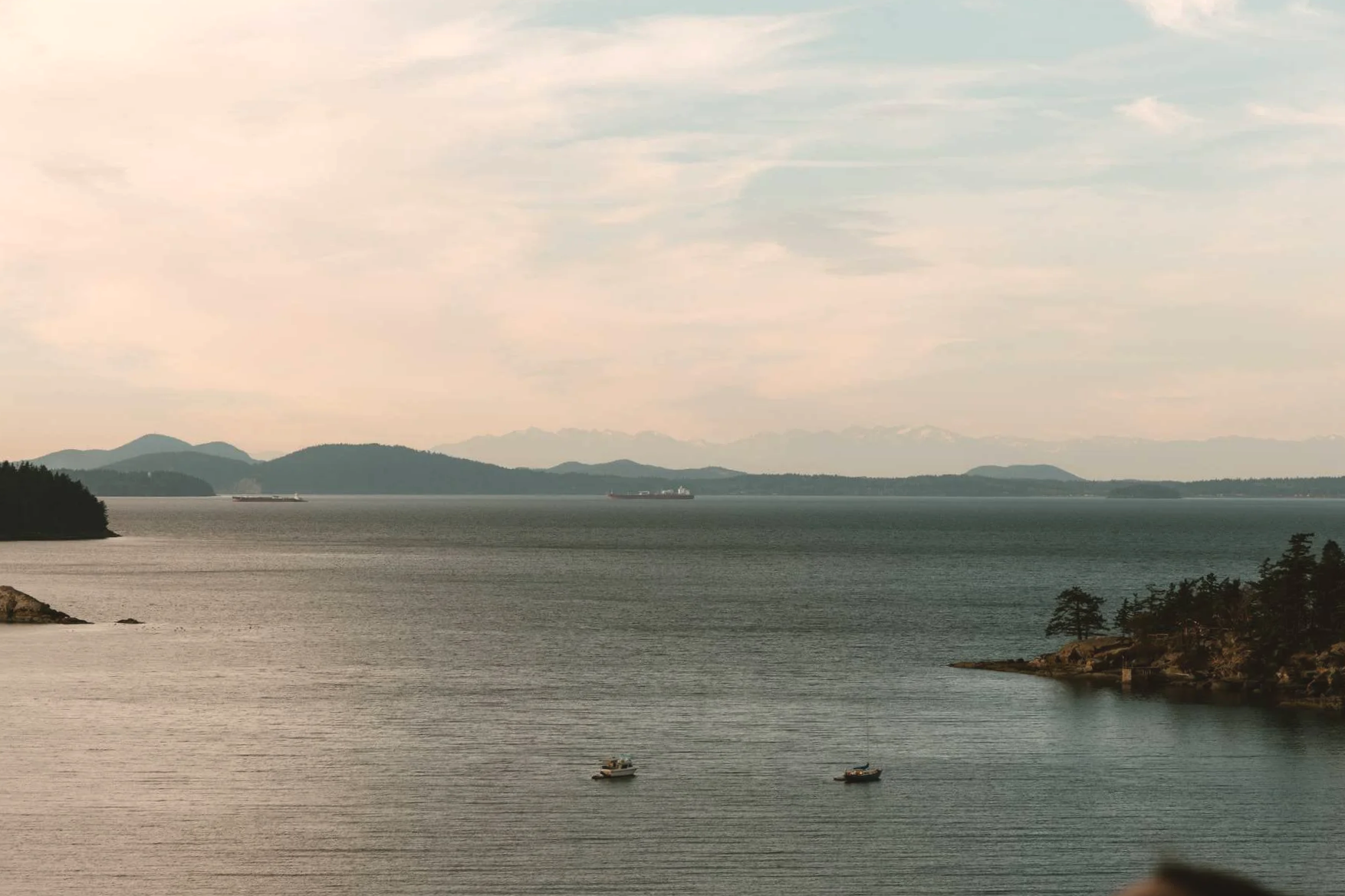 A scenic view of a large body of water with two small boats in the foreground, distant islands and mountains in the background, and a cloudy sky overhead.