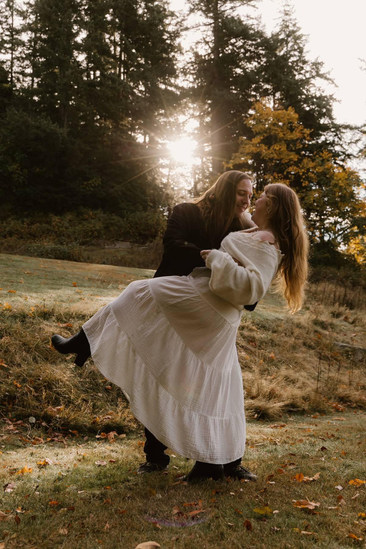 Two women sharing a joyful moment outdoors, one lifting the other in her arms with trees and the setting sun in the background.