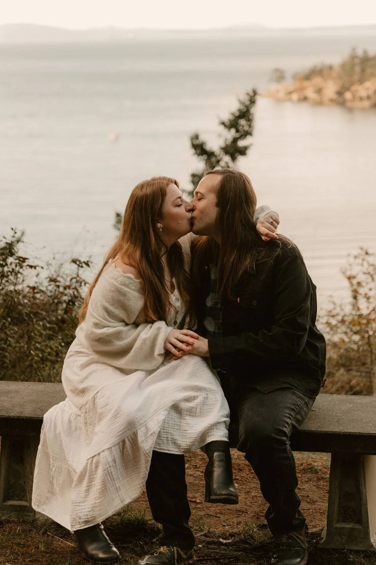 A couple sharing a kiss on a bench near a lake, surrounded by trees with fall foliage.