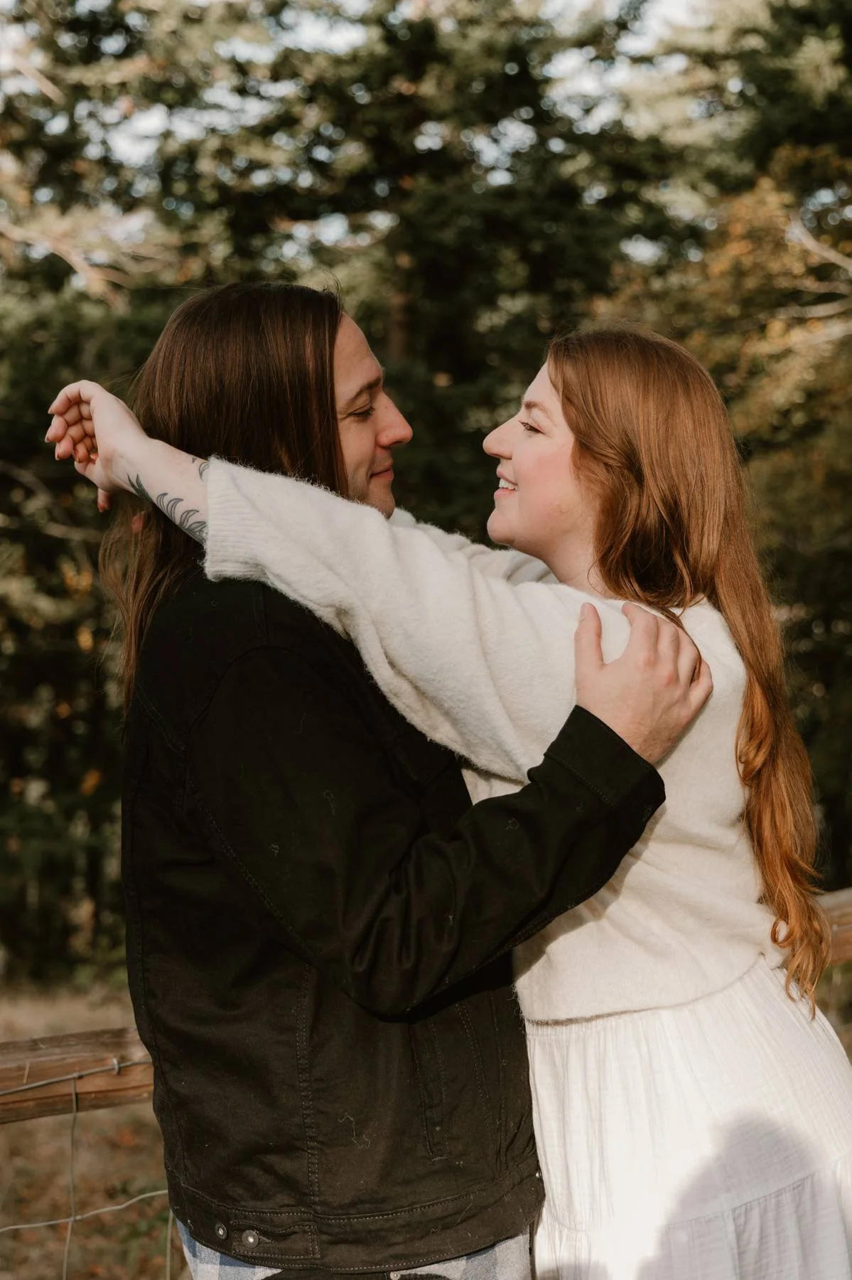 A couple embraced outdoors, smiling into each other's eyes with trees in the background.