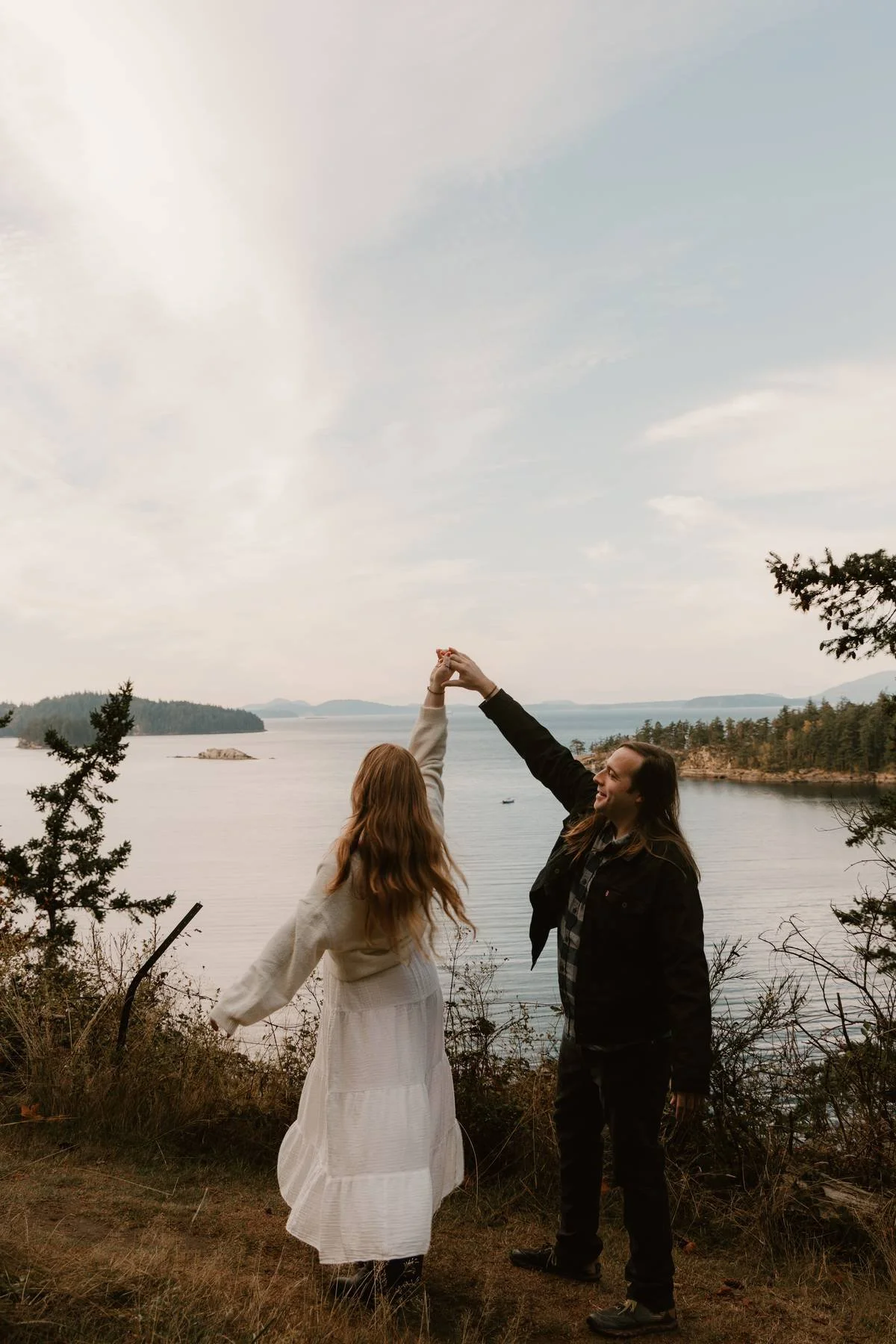 A man and a woman dancing together outdoors near a lake with trees and mountains in the background.