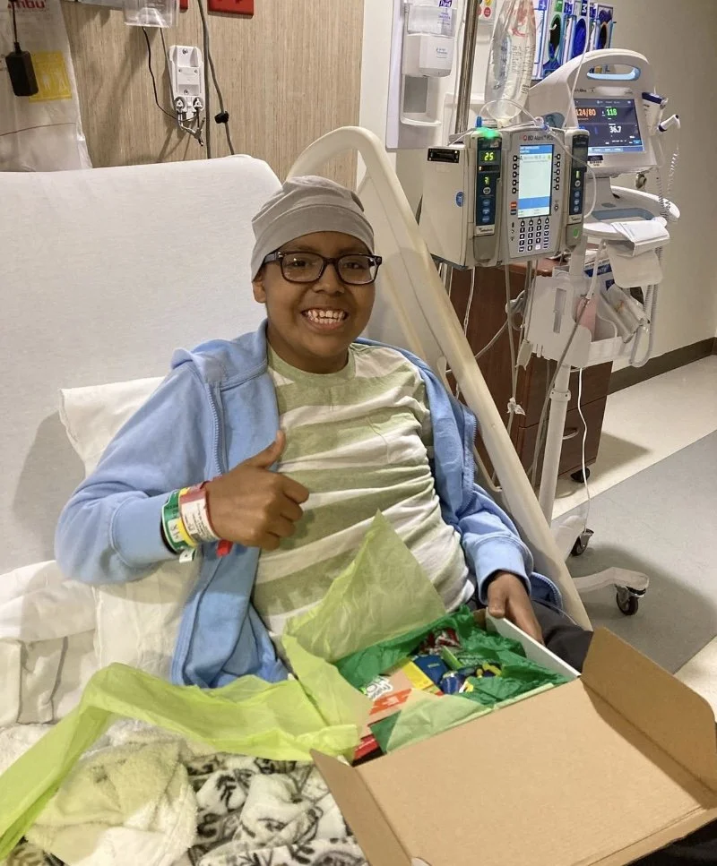 Child in a hospital bed smiling and giving a thumbs-up while holding a box of snacks, with medical equipment behind.