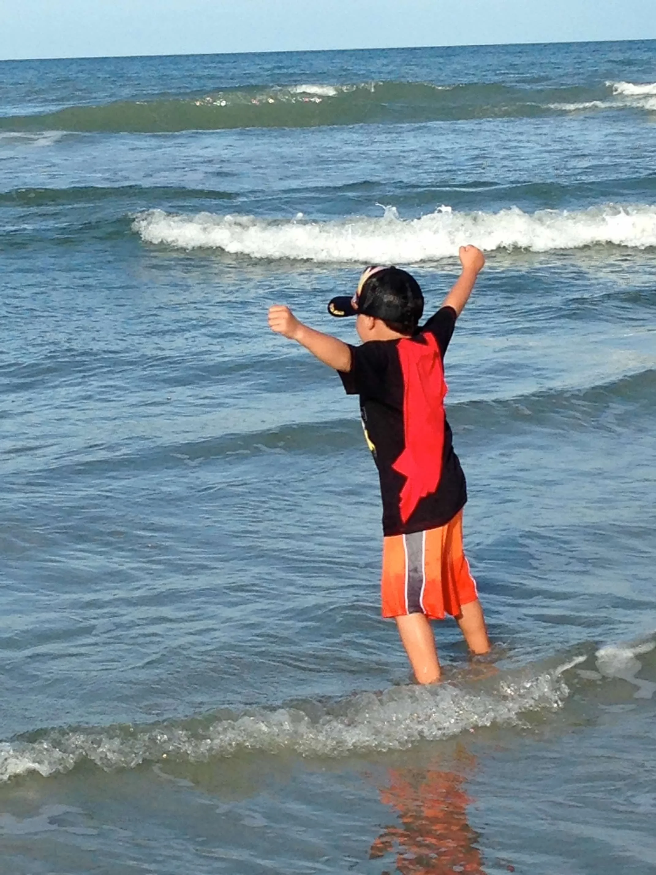 A young boy wearing a black cap, red life jacket, and orange shorts is standing in shallow ocean water at the beach with his arms raised, facing the waves.