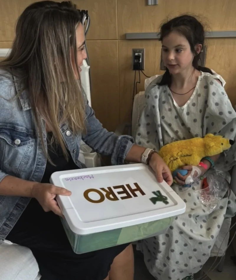 Young girl in hospital gown sitting in hospital bed receiving a hospital care package from a woman, likely her mother, in a denim jacket, who is holding a plastic container labeled 'HERO' with a printed Design of a cacti and the word 'Macaroni.'