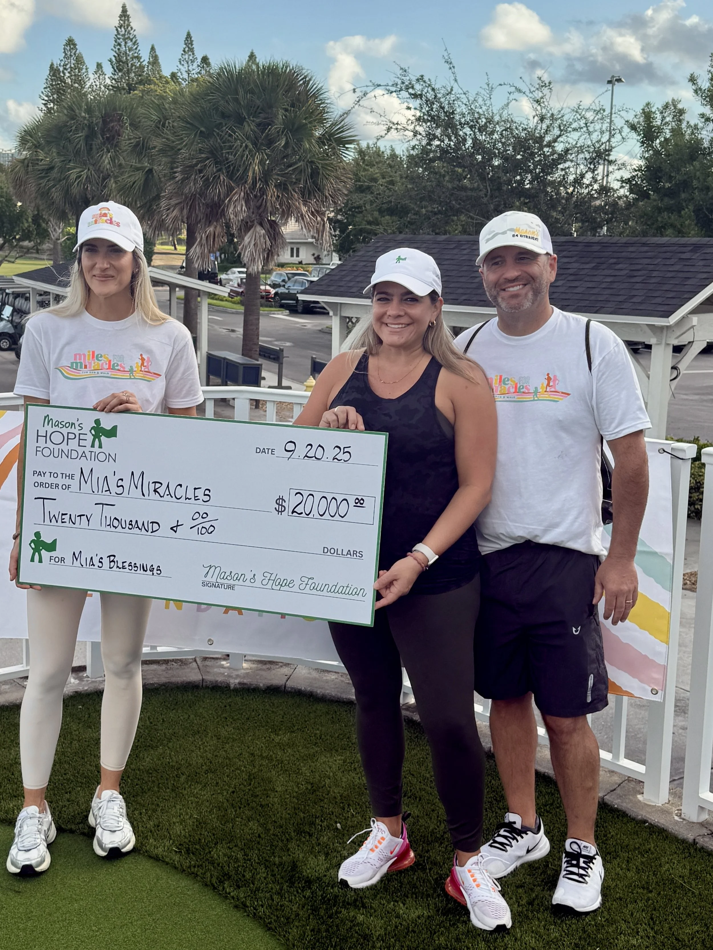 Three people standing together outdoors, holding a large check for twenty thousand dollars made out to Mia's Miracles, dated 9.20.25, at Mason's Hope Foundation event, with smile and casual athletic wear. The background features trees, a parking lot, and a partly cloudy sky.