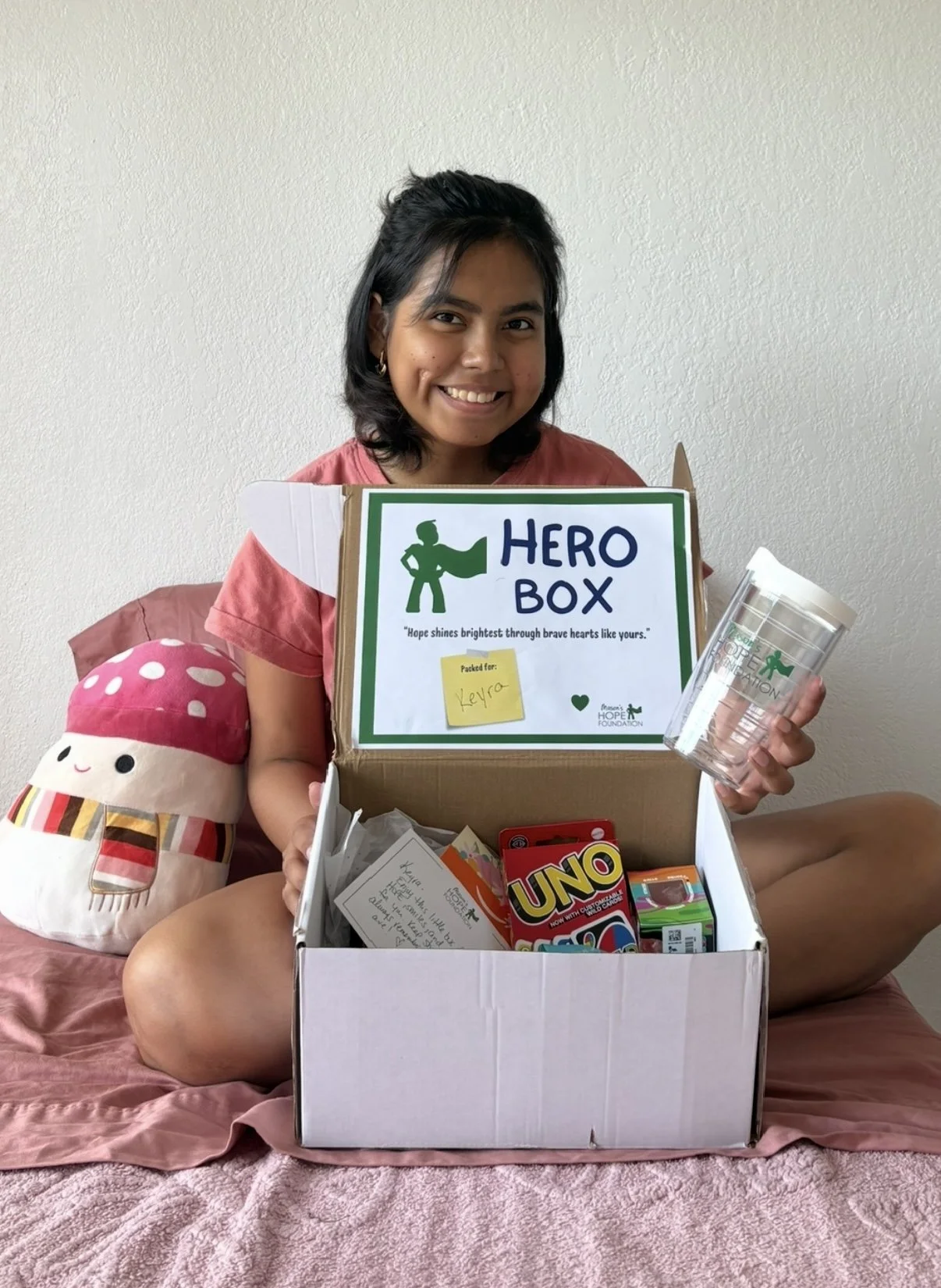 Young woman sitting on a bed holding a box labeled 'Hero Box' filled with toys and supplies, with a plush mushroom-shaped stuffed toy nearby, and smiling at the camera.