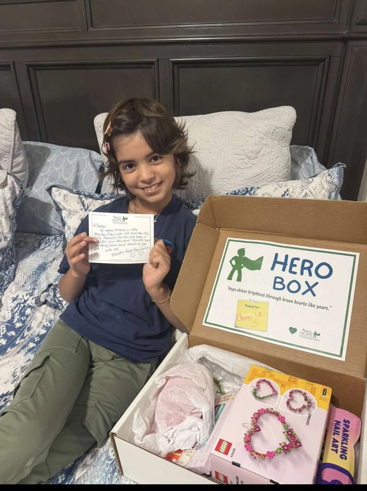 A girl sitting on a bed smiling and holding a handwritten note, with an open box labeled 'Hero Box' beside her. The box contains craft supplies, including a jewelry making kit and tissue paper. The girl has dark hair with hair clips and is wearing a navy shirt and light-colored pants.