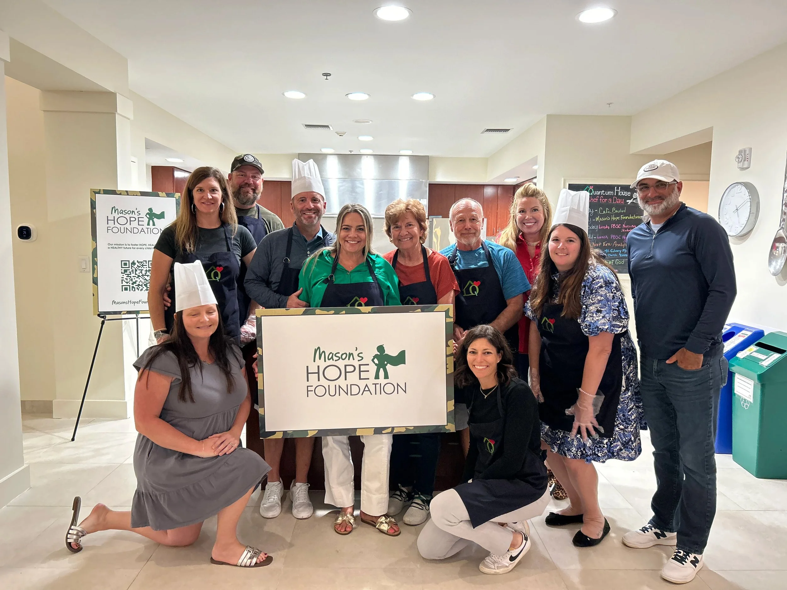 Group of people standing behind a Mason's Hope Foundation sign, smiling, in a brightly lit indoor space. Some are wearing aprons and chef hats, indicating a cooking event.