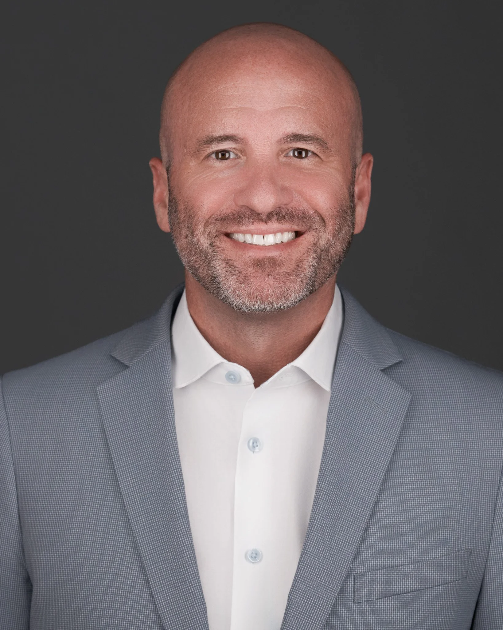 Headshot of a bald man with a beard, smiling, wearing a light gray suit and white shirt, against a dark background.