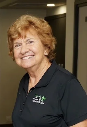 A smiling woman with short, curly red hair wearing a black polo shirt with the Hope Foundation logo.