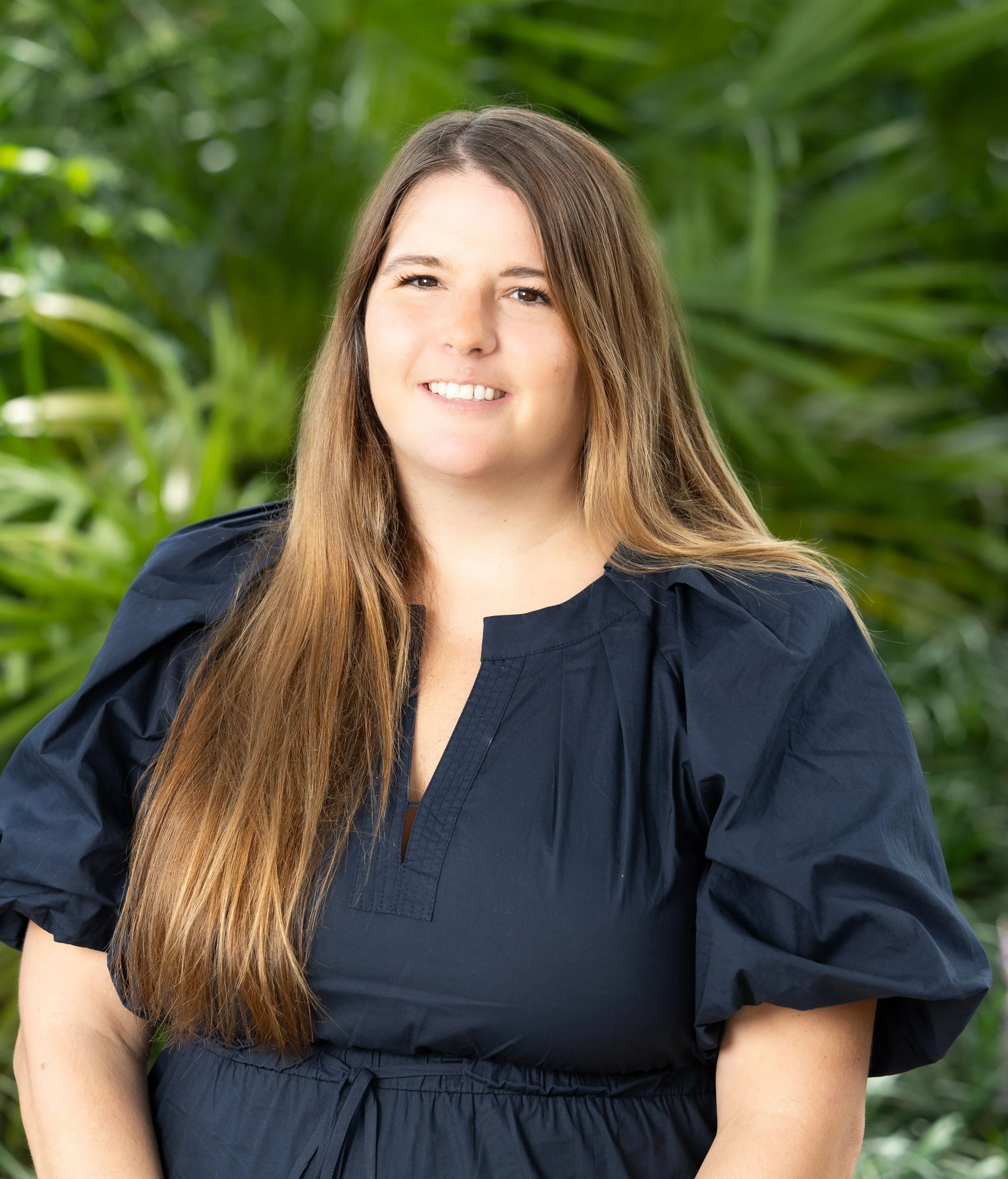 Close-up of a woman with long brown hair, smiling, wearing a navy blue top, outdoors with a background of green foliage.