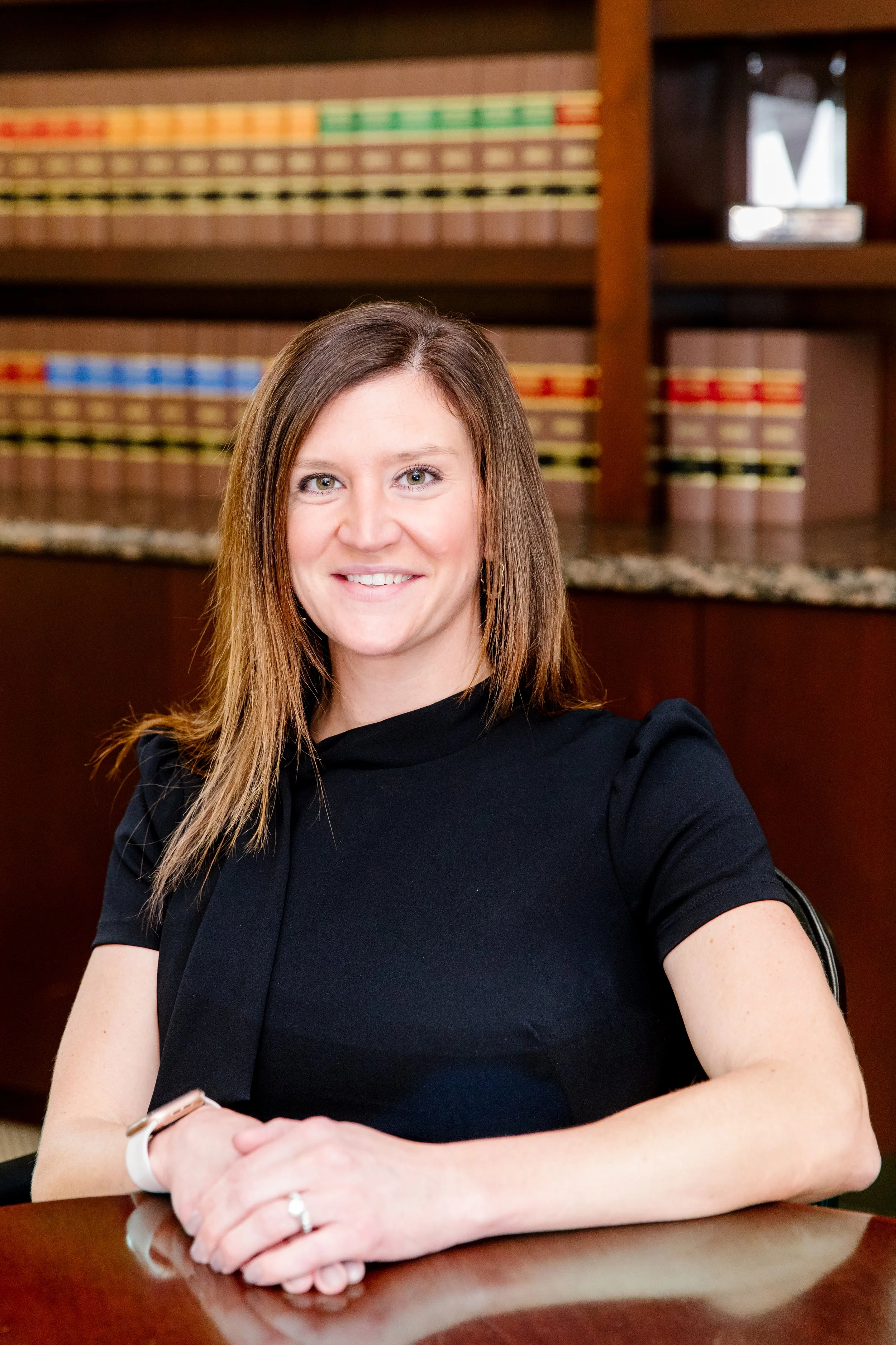 A woman with shoulder-length brown hair, wearing a black dress, smiling and sitting at a wooden table in a room with bookshelves filled with legal books in the background.
