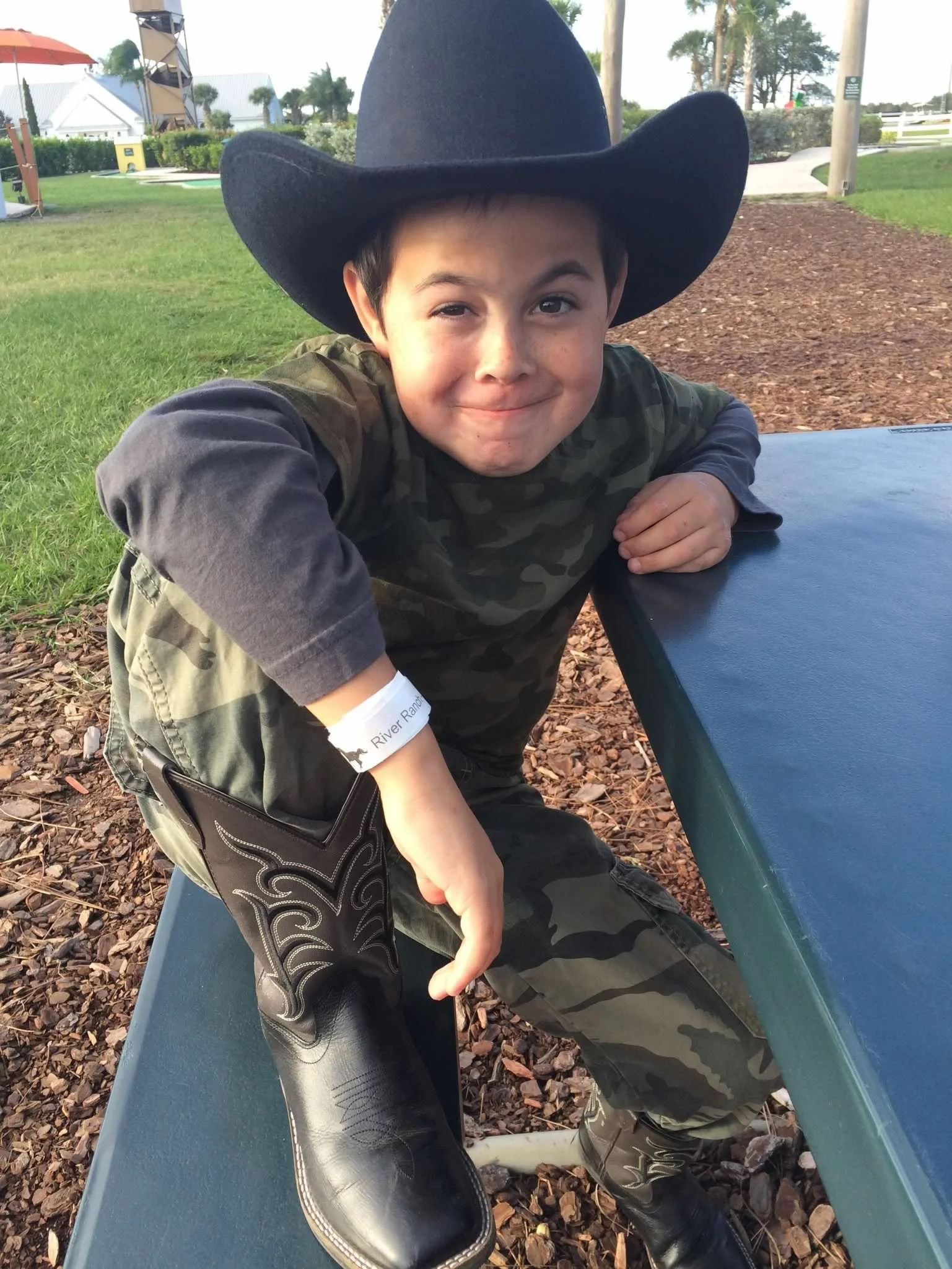 A young boy wearing a large black cowboy hat and camouflage clothing is leaning on a blue outdoor table, smiling towards the camera. There is green grass, trees, and structures in the background.