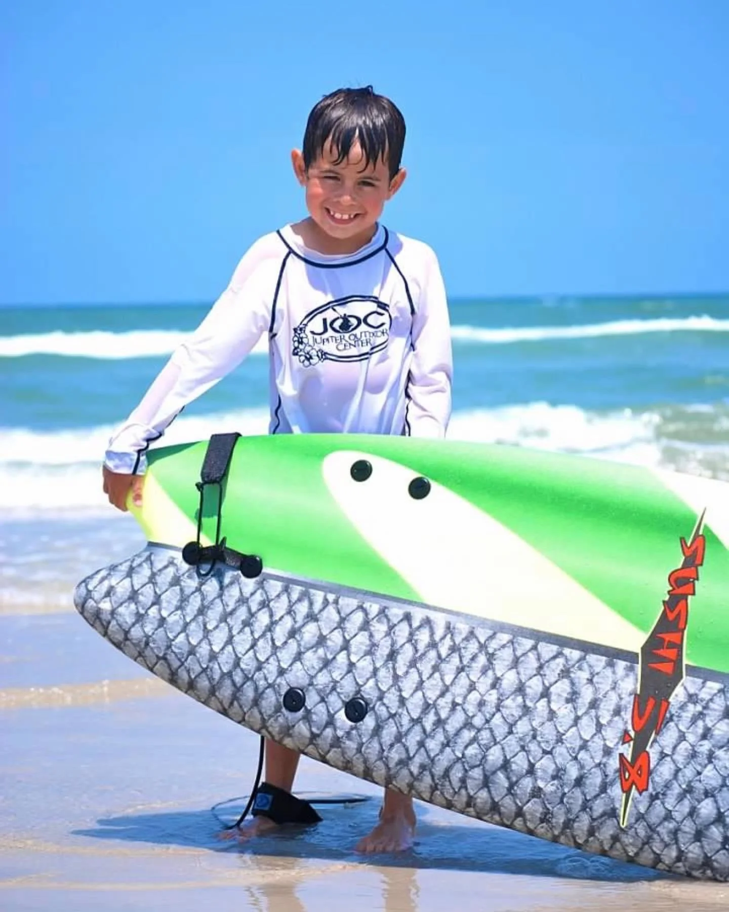 A young boy smiling and holding a surfboard at the beach with ocean waves in the background.