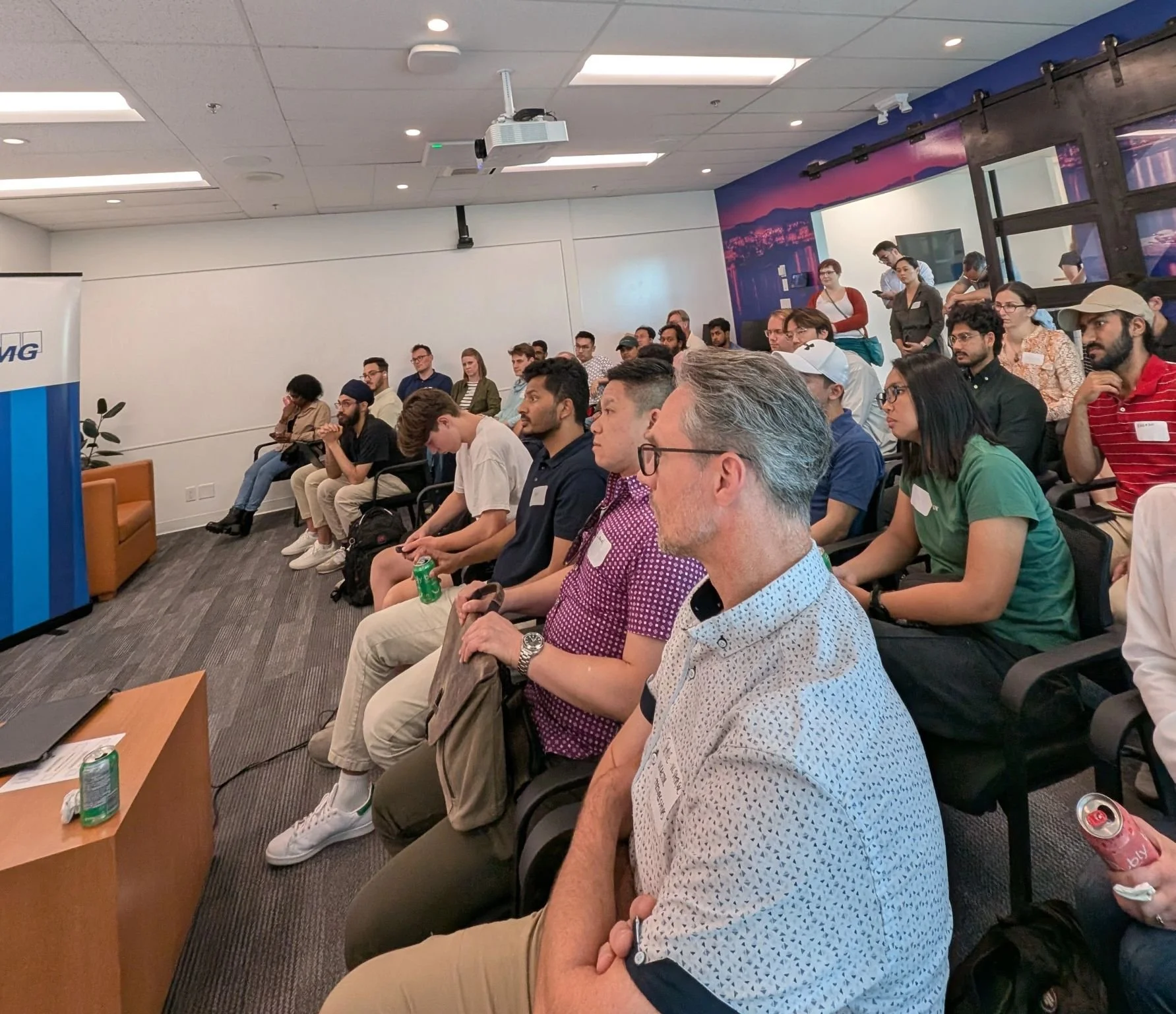 An audience sitting in a conference room, attentively listening to a presentation. There is a window with a view of city buildings. Some people are taking notes, and there are drinks on the desks.