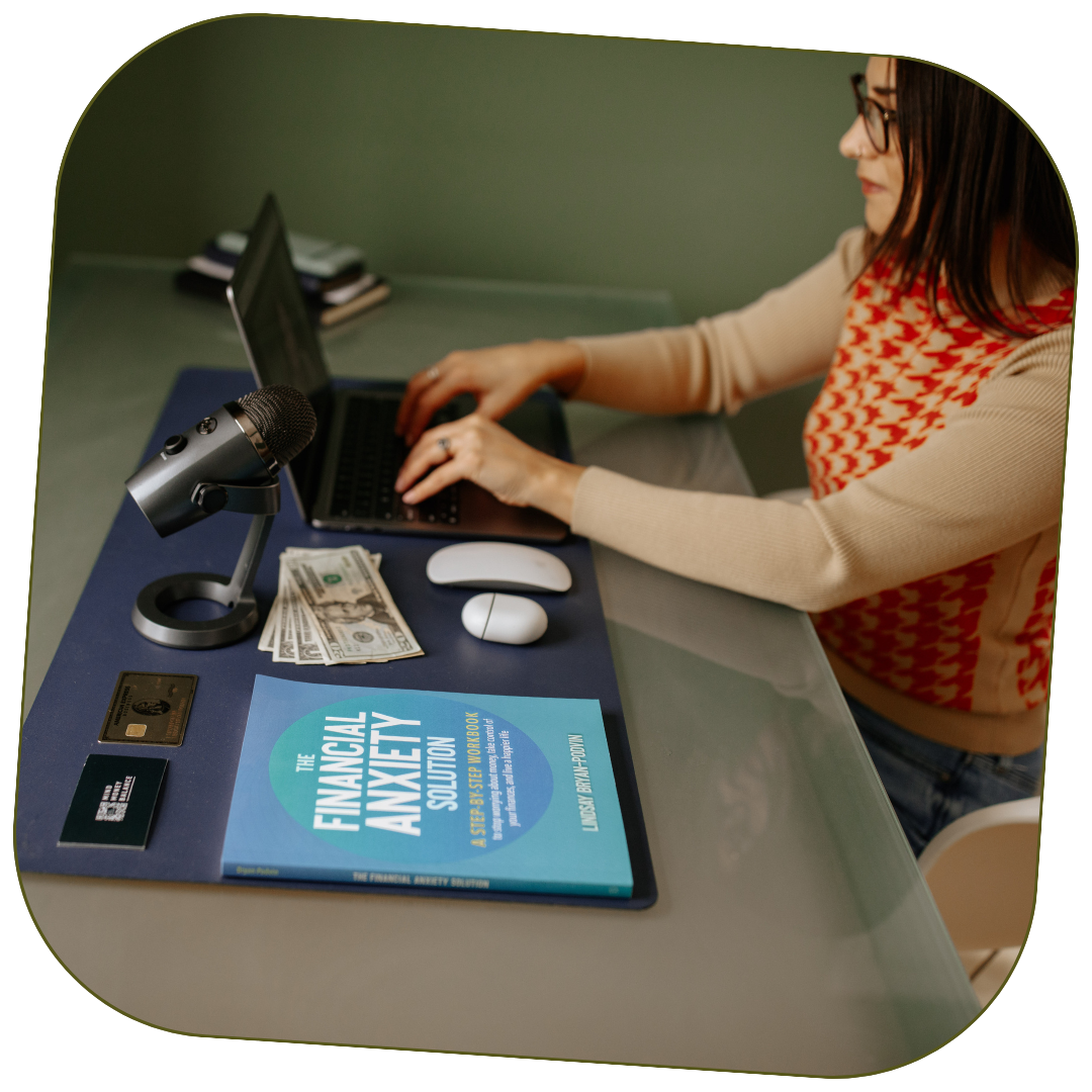 Founder Lindsay Bryan-Podvin is sitting at a desk with a laptop, microphone, cash, credit cards, a book titled 'The Financial Anxiety Solution', and a computer mouse. She's finalizing information on the Mind Money Balance Method.