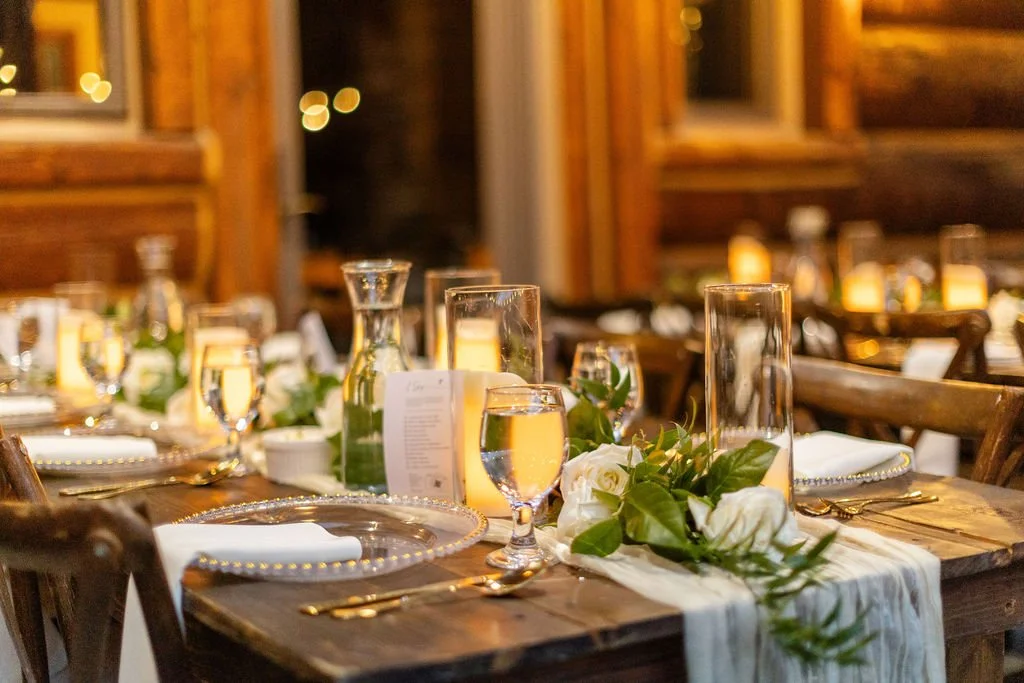 Close up of table setting with whit table runners, green garland, white roses, gold beaded chargers, gold flatware, and ivory napkins