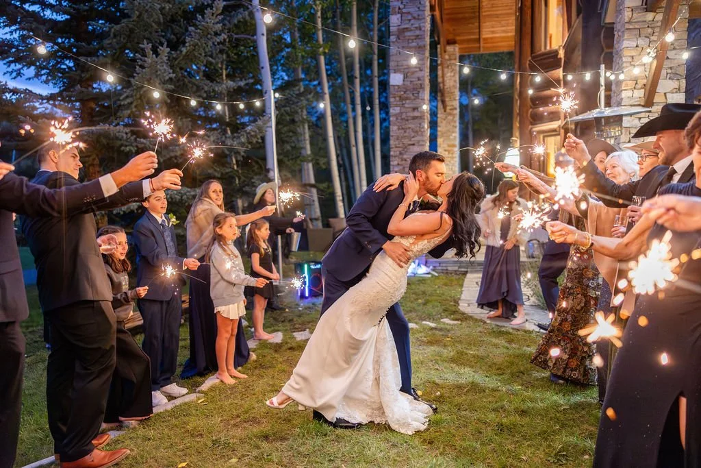 Bride and groom kiss under string lights while guests wave sparklers