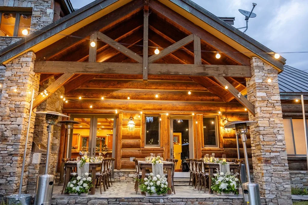 Farm tables set up on back patio of brick home with bistro lights and floral arrangements
