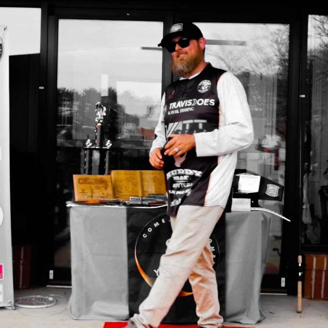 A man with a beard wearing sunglasses, a black and white cycling jersey, and light-colored pants standing in front of a table with trophies and awards.