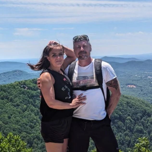 A man and woman standing together outdoors on a mountain, with green hills and a cloudy sky in the background.